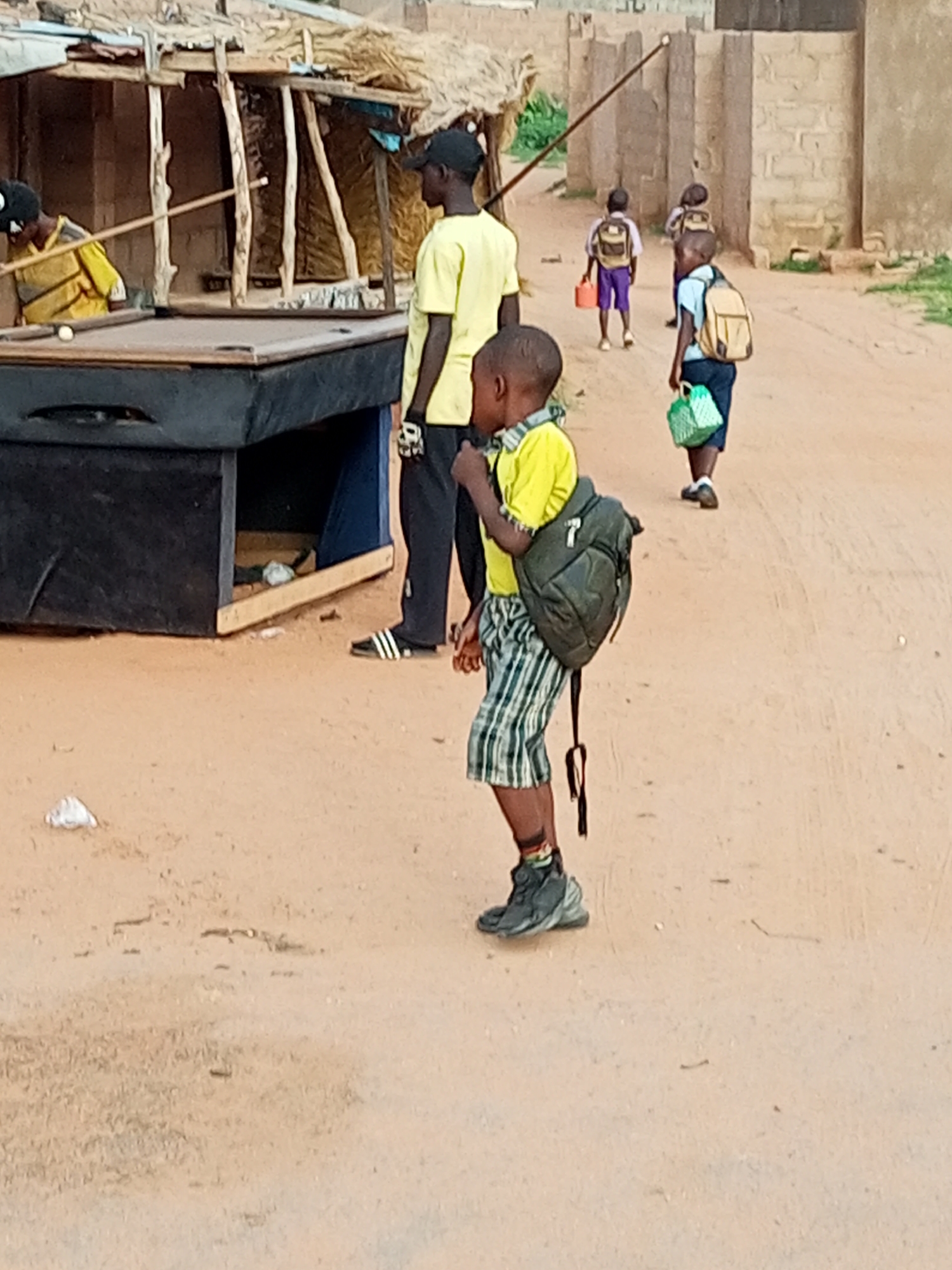 In Tumfure, Nigeria, on a dirt road, three young boys and two older individuals are visible. The older individuals are an adult male wearing a black cap and a yellow t-shirt, standing near a pool table, and another adult male wearing a black t-shirt. One of the young boys, wearing a yellow t-shirt and plaid shorts, has a dark green backpack and is walking to the right. Another young boy in a light blue t-shirt and dark shorts also has a backpack and is carrying a green patterned bag. A third young boy, wearing purple shorts and a light colored t-shirt, carries a red lunchbox. In the background, there are simple structures with thatched roofs and brick buildings. The scene suggests a community setting, possibly near a school or recreational area, with children preparing to go somewhere.