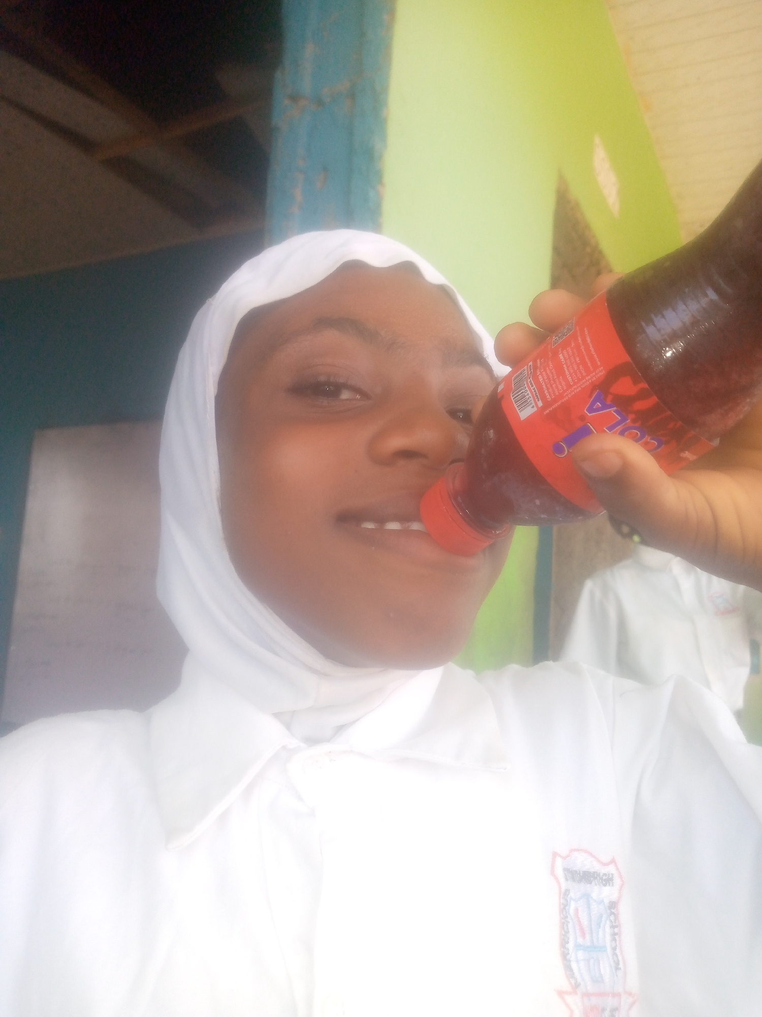 A person with brown skin is wearing a white hijab and a white shirt with an emblem on the left side. The person is holding a bottle of cola and tilting it towards their mouth. The bottle has a red label and a red cap. The cola appears to be red-brown in color. The background shows a combination of dark and light green walls and what appears to be a window or a door with blue trim. Another person wearing white is partially visible in the background. The location is Unknown, Unknown.
