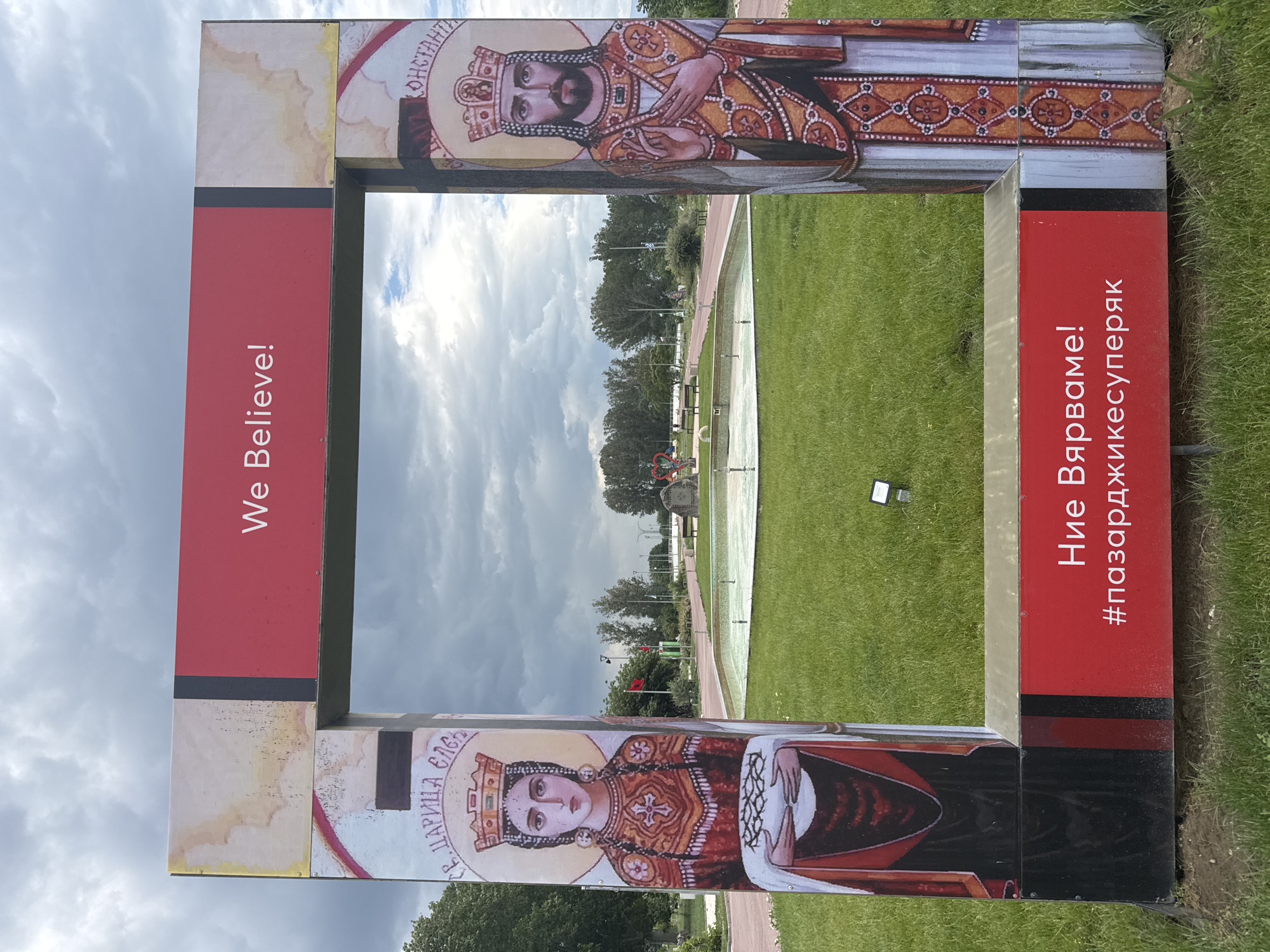 A ground-level, outdoor, medium shot captures a large, decorative frame in the city of Unknown, Unknown, with artwork and text on its four sides, framing a green park landscape beyond. The frame has two distinct images: at the top, an Orthodox Christian icon of a male figure, probably Constantine the Great, wearing a crown and ornate robes; at the bottom, a similar icon of a female figure, possibly Saint Helena, also with a crown and robe. The figures appear to be painted or printed reproductions.
The left side of the frame features the English text "We Believe!" printed vertically on a red background. The right side displays similar text in a different language, possibly Bulgarian, and the hashtag "#пазарджиксуперяк" printed on a red background. The frame itself is constructed from panels, suggesting a temporary or art installation.
The park landscape through the frame consists of a grassy area, walkways, and a long, shallow water feature with small fountains. Trees are visible in the background, adding depth to the scene. The sky above is overcast with clouds, indicating a potentially cloudy day. A small, metallic object, possibly a ground light or fixture, is visible on the grass to the right. The overall setting implies a public space or park area with elements of religious or historical commemoration.