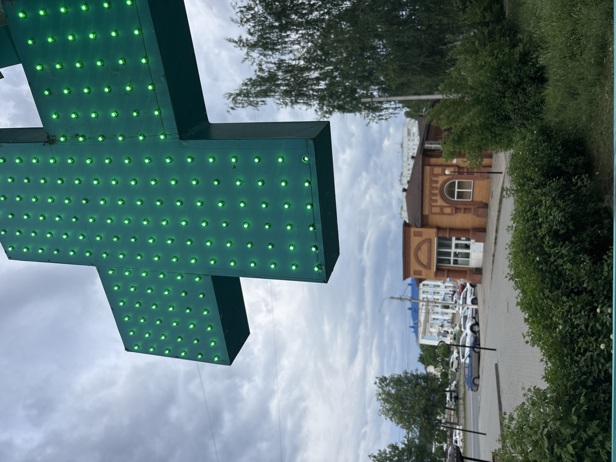 The image shows a street scene in the city of Chaikovsky, Russia. A large green illuminated cross, likely a pharmacy sign, dominates the foreground. Behind it, there is a view of a street with parked cars, buildings, trees, and cloudy sky. The architecture includes a light-brown brick building with arched windows. Foliage is visible along the side of the street. The scene is captured from an oblique angle.
