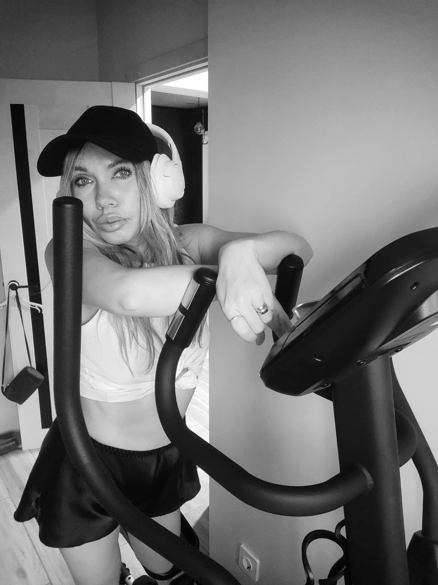 A young woman is leaning on an elliptical machine. She is wearing a black baseball cap, white headphones, a white crop top, and black shorts. A small purse is hanging from a door frame in the background. The image is in black and white. It is not possible to verify the location in the city of Votkinsk, Russia from this image.