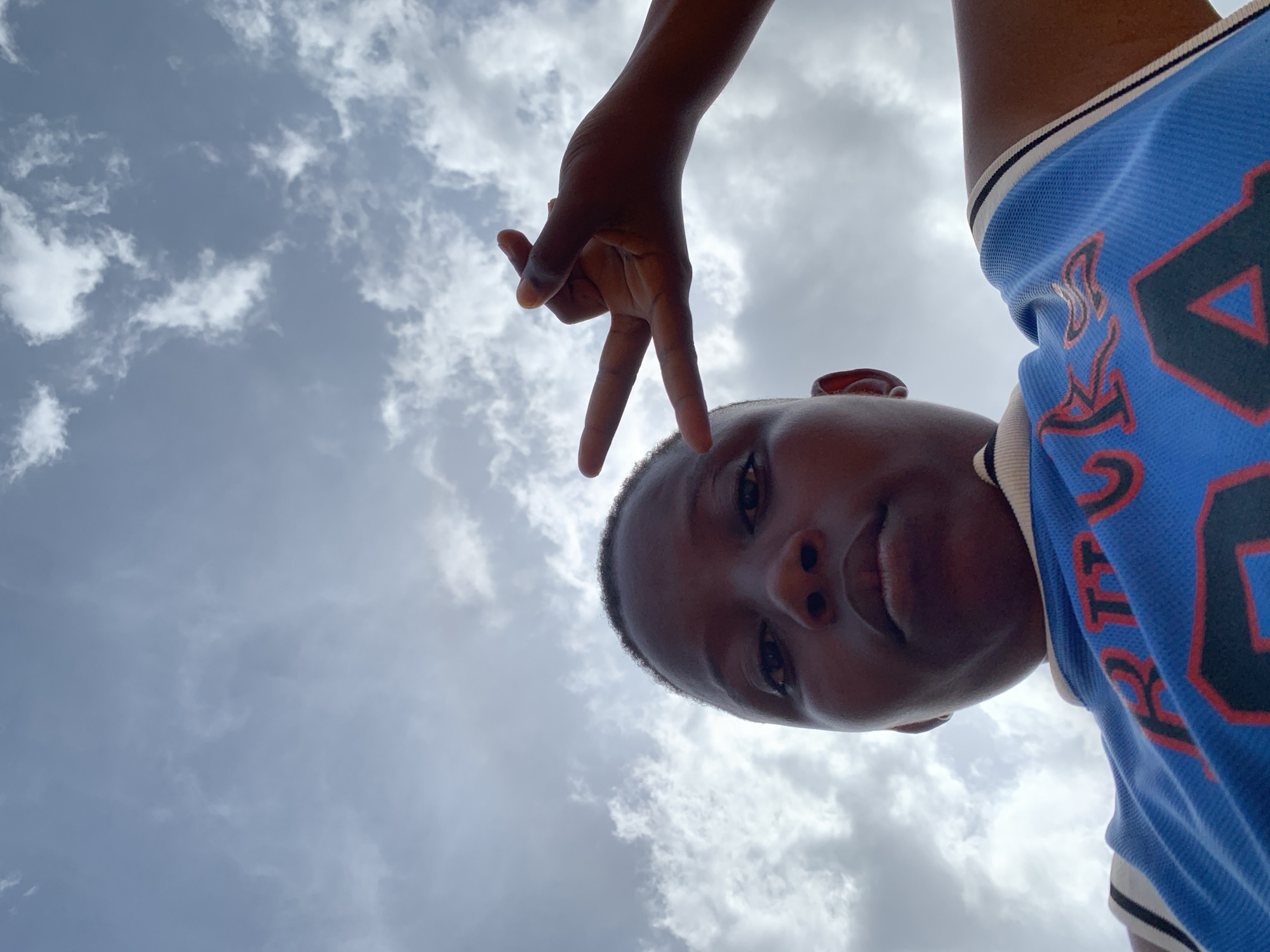 A low-angle shot shows a dark-skinned person posing against a cloudy sky. The person is looking down at the camera and making a peace sign with their right hand, which is positioned above their head. The individual is wearing a blue basketball jersey with white and black trim around the neck and shoulders, and what appears to be a large black number is visible on the jersey. The location is not discernible as being in the city of Ikorodu, Nigeria, as the environment appears to be only sky and the subject.