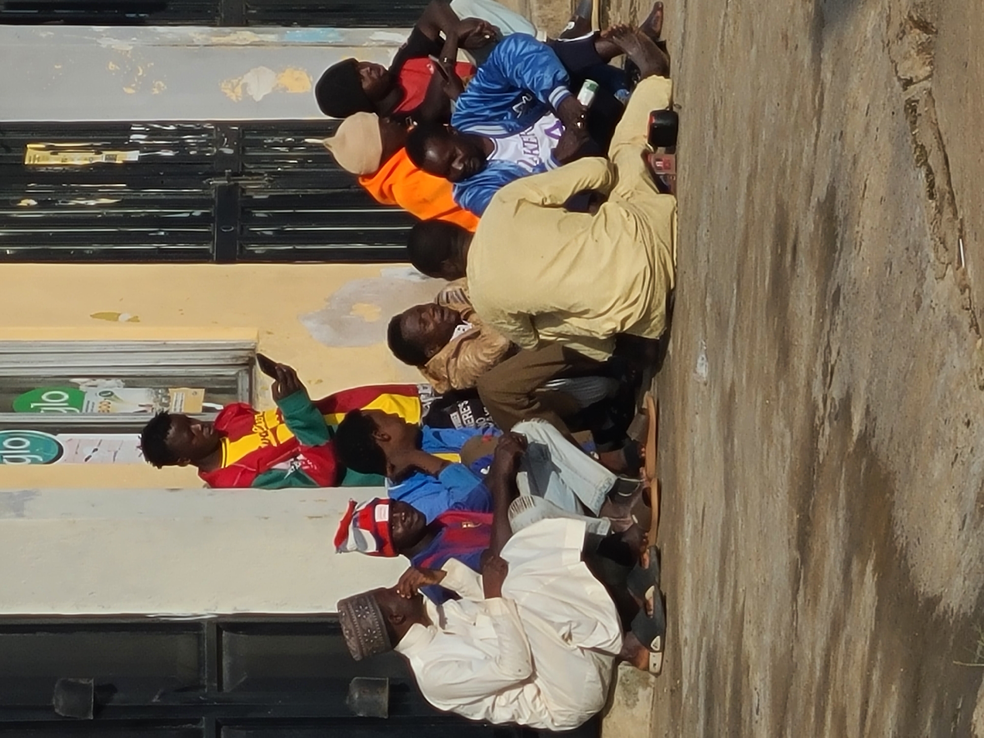 A group of nine Nigerian men are gathered outdoors in front of a building with a black, barred door and a light yellow, partially peeling wall. The men are of varying ages and are dressed in casual attire, including traditional clothing and modern sportswear. Some are sitting on the ground, while others are seated on a ledge. One man, wearing a white traditional robe and cap, is seated on the left. Next to him, two men are seated wearing a striped jersey and a blue jacket respectively. Further to the right, a man in an orange hoodie and another in a black t-shirt are standing. In the foreground, a man in a beige traditional outfit is seated on the ground. Another man in a blue jacket is seated next to him, looking at his phone. Two other men are seated in the background. The ground appears to be concrete and wet in places. The scene suggests a casual gathering or waiting area in an urban environment. The presence of "Glo" signage on the building suggests this location may be near a telecommunications provider's outlet. The overall atmosphere is relaxed and informal.