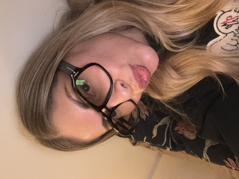 A woman with blonde hair poses in a sideways, close-up shot. She wears dark-rimmed glasses and has on a dark blouse with floral designs. An embroidered patch with a white background and red and black details is partially visible. Her lips are slightly pursed. The background appears to be a plain, light-colored surface.