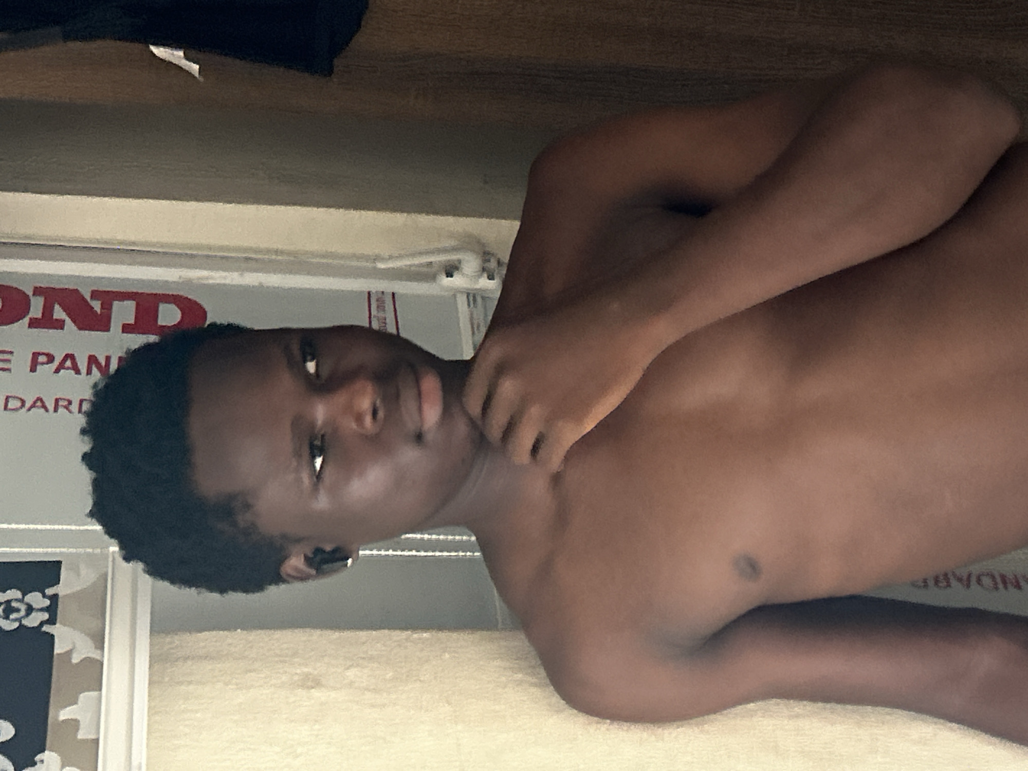 A shirtless dark-skinned young man with short, dark hair is lying on a white surface. His right arm is bent and positioned near his neck. He is looking directly at the camera. Behind him is a wall or panel with red lettering "OND E PAN STANDARD". There are floral decorations on a dark background to the left of the wall. The environment suggests an interior space. There is no explicit information to indicate a specific location in the city of Ihiagwa, Nigeria.