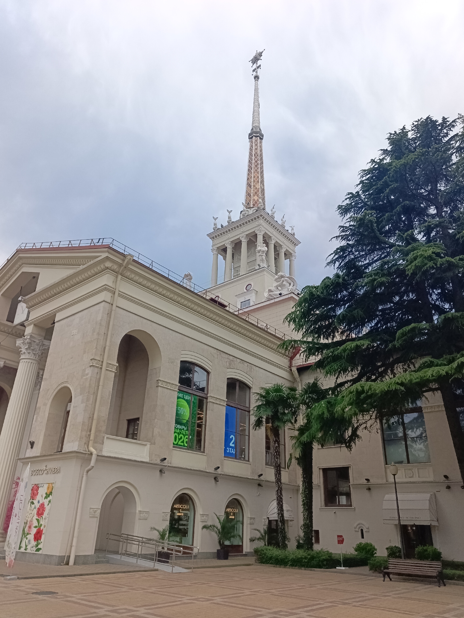 The image shows the Sochi Sea Port building in Sochi, Russia, on a cloudy day. The port's distinctive white facade with arched windows and columns is visible, as is its tall, ornate spire topped with a sculpture. Large evergreen trees and palm trees are present, adding to the tropical atmosphere of the city. The ground in front of the building is paved with a checkered pattern. Signage on the building indicates shops, including "BOSCO RIVIERA" and "ARTICOLI". The scene is devoid of people.