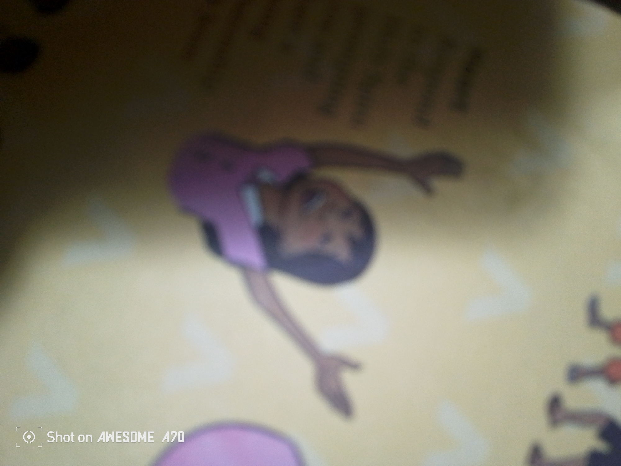 The image is a blurry, low-resolution photograph of what appears to be a page from a book or comic. A stylized illustration of a person in a pink top and dark bottoms is the central focus. The person is depicted in a dynamic pose, possibly a dance or acrobatic move, with their body arched backward and arms extended. Their face is visible, with dark hair and brown skin. The background is a pale yellow, with some indistinct shapes and text. In the bottom right corner, there are blurred figures that appear to be other people, possibly in a line, wearing orange tops and dark bottoms, and some are holding red objects. There is a watermark in the bottom left corner that reads "Shot on AWESOME A70". The image does not contain any information or visual cues that would allow for a determination of its location in Jalingo, Nigeria.