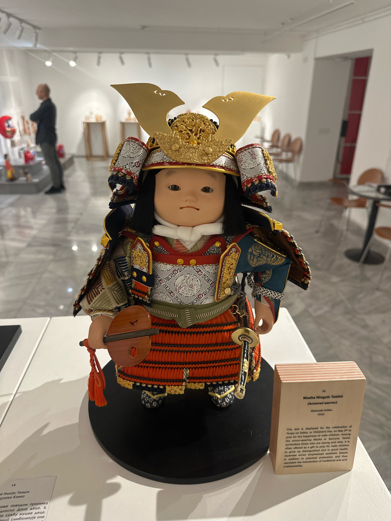 A detailed view of a Japanese warrior doll, identified as "Musha Ningyō: Taishō (Armored warrior)" by Matsuzaki Koiko, 2020, is presented. The doll is adorned in elaborate samurai armor with gold accents, intricate fabric patterns, and a distinctive helmet. It stands on a black circular base. To the right of the doll, a small wooden plaque provides information about the artwork. The doll is displayed in a museum or gallery setting, indicated by the white walls, track lighting, and polished floor. In the background, blurred figures of people and display cases suggest an exhibition environment. The overall lighting is bright and even, illuminating the doll and its immediate surroundings. The image appears to have been taken in the Museum of Yugoslavia in Belgrade, Serbia, given the context of displayed cultural artifacts.