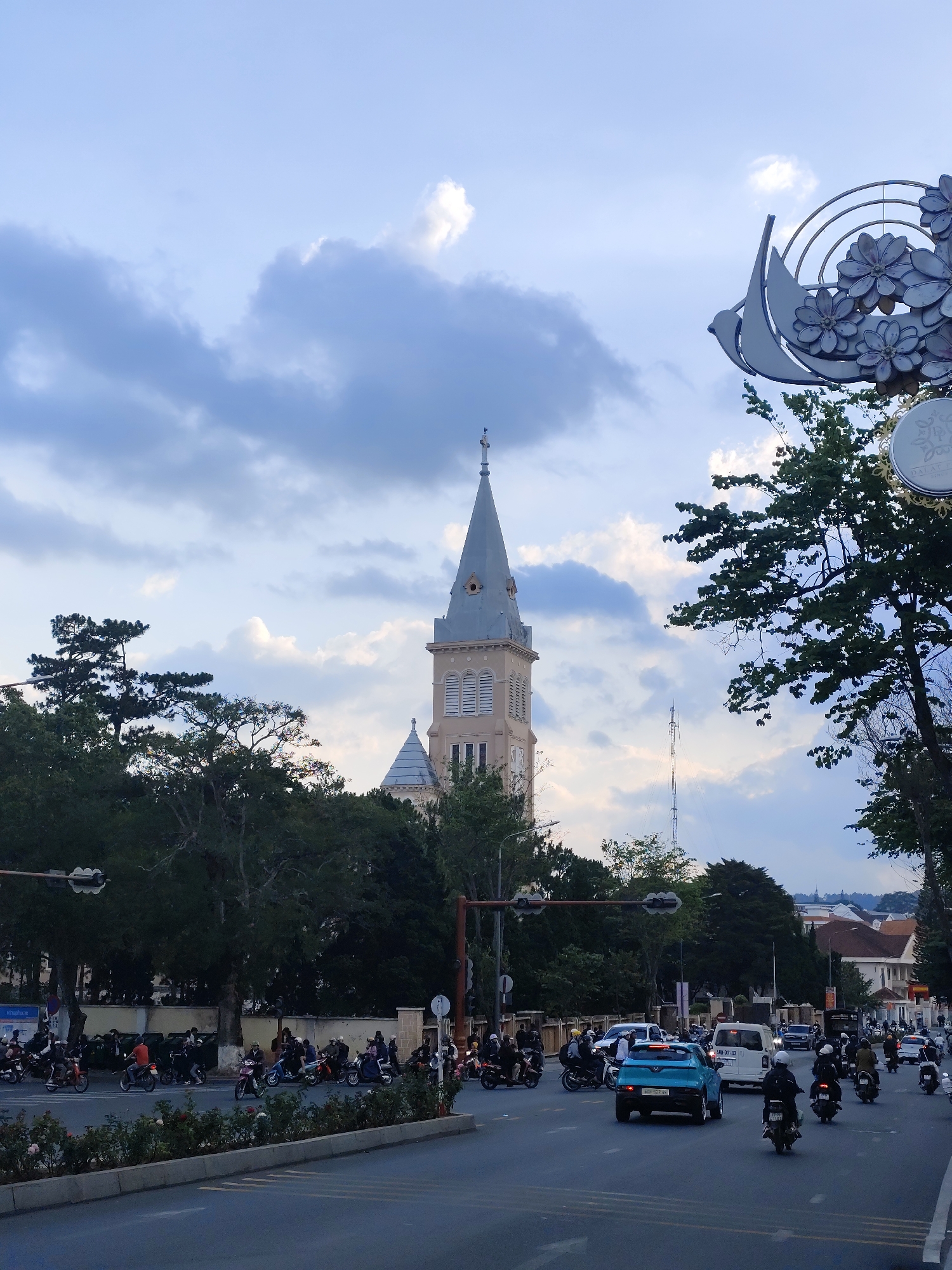 The image depicts a street scene in Da Lat, Vietnam, with the Da Lat Cathedral (also known as the Chicken Church) prominently visible in the background. The sky is overcast with clouds. A busy road with motorcycle traffic is in the foreground. There are trees lining the side of the road and a decorative street sign with a bird and flower design in the upper right corner. Cars and a white van are mixed in with the motorcycles on the road. The photograph appears to have been taken during the daytime.