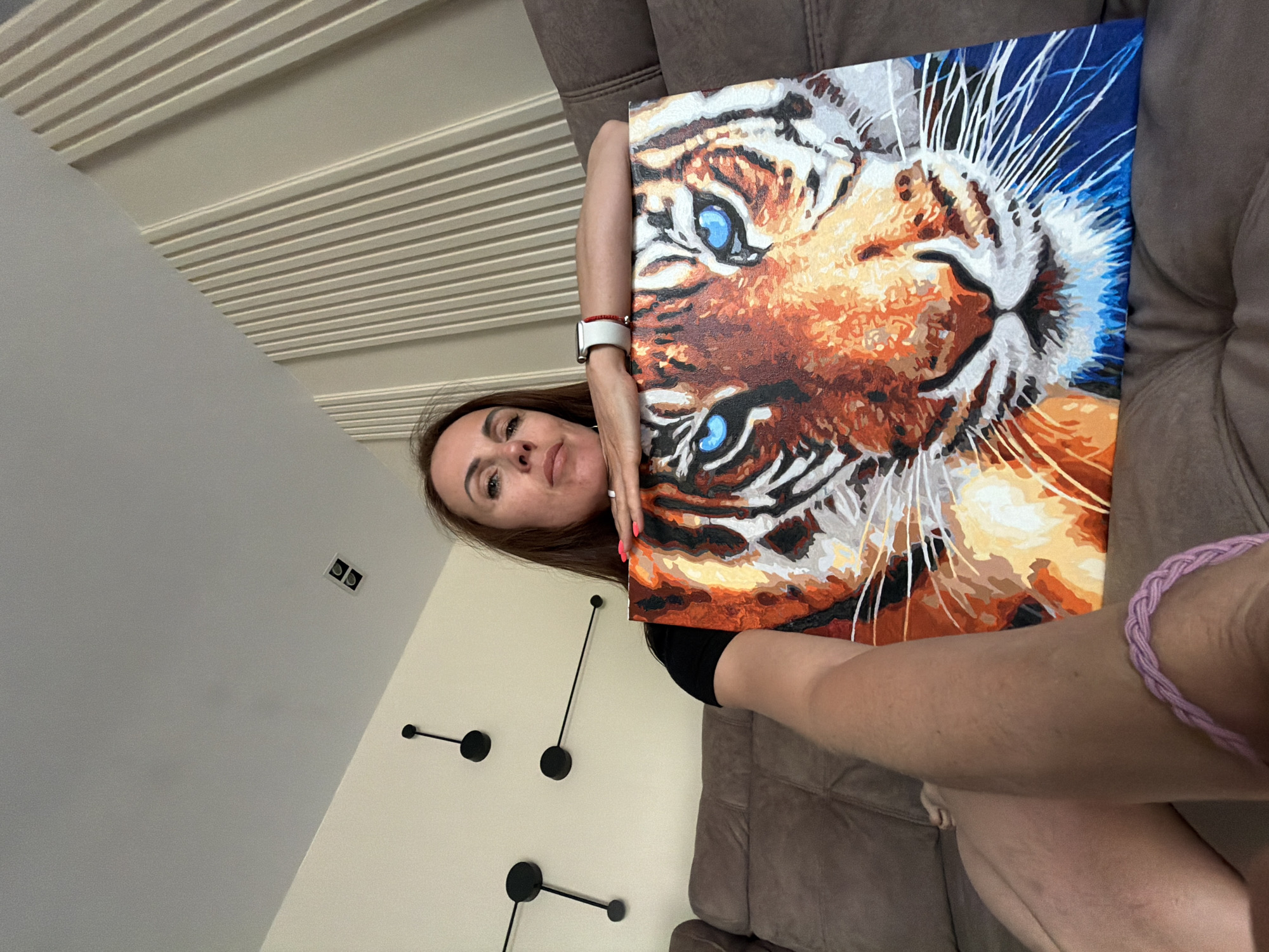 A woman is pictured indoors, seated or leaning against a brown upholstered sofa, proudly displaying a painting. She holds a vibrant artwork depicting a tiger's head, which features bold orange, white, and black stripes, and striking blue eyes. The woman has long brown hair and looks directly at the camera with a subtle, content smile. She wears a dark top, a white smartwatch with a red band on her left wrist, and a light pink braided bracelet on her right wrist.
The background consists of a light-colored wall adorned with three modern, minimalist decorative elements that resemble parts of a clock, featuring black circular pieces connected by thin black rods. Above the wall, a ceiling with distinct horizontal white slats is visible, along with a couple of small, rectangular recessed fixtures in the upper left corner. The lighting suggests it is daytime. The overall scene conveys a sense of personal achievement or pride as the woman showcases her artwork in what appears to be a comfortable domestic setting. There is no visible text in the image.