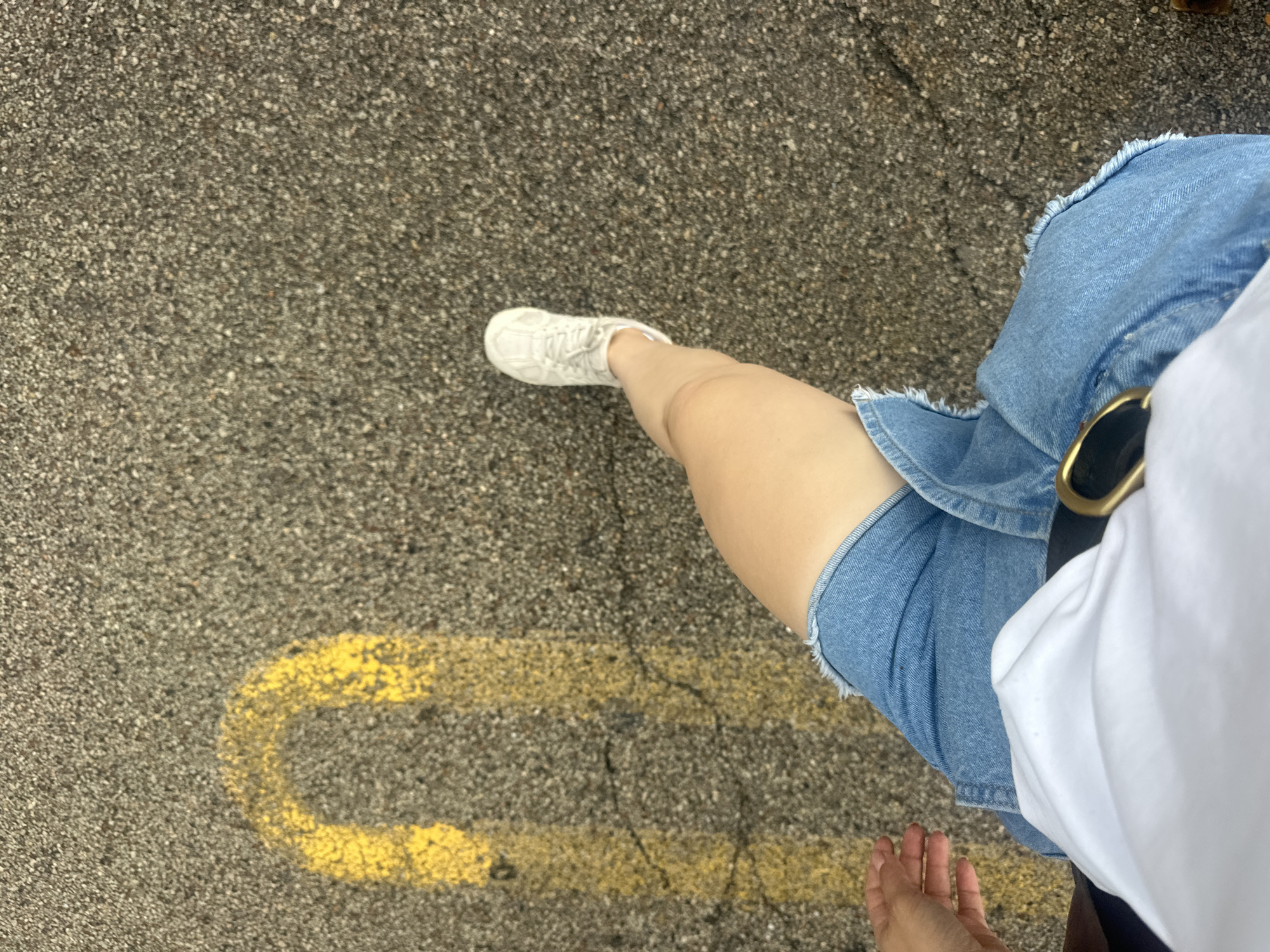 The image is a first-person perspective shot of a person walking on asphalt. The person is wearing a white t-shirt, a denim skirt with a frayed hem, and white sneakers. A gold belt buckle is visible on the skirt. Yellow markings, possibly from parking lines or road markings, are present on the asphalt, including a U-shaped line and several parallel lines. The ground is cracked in places. The setting appears to be an outdoor urban environment, consistent with locations in Schaumburg, United States of America, such as a parking lot or a street. The lighting suggests it is daytime.