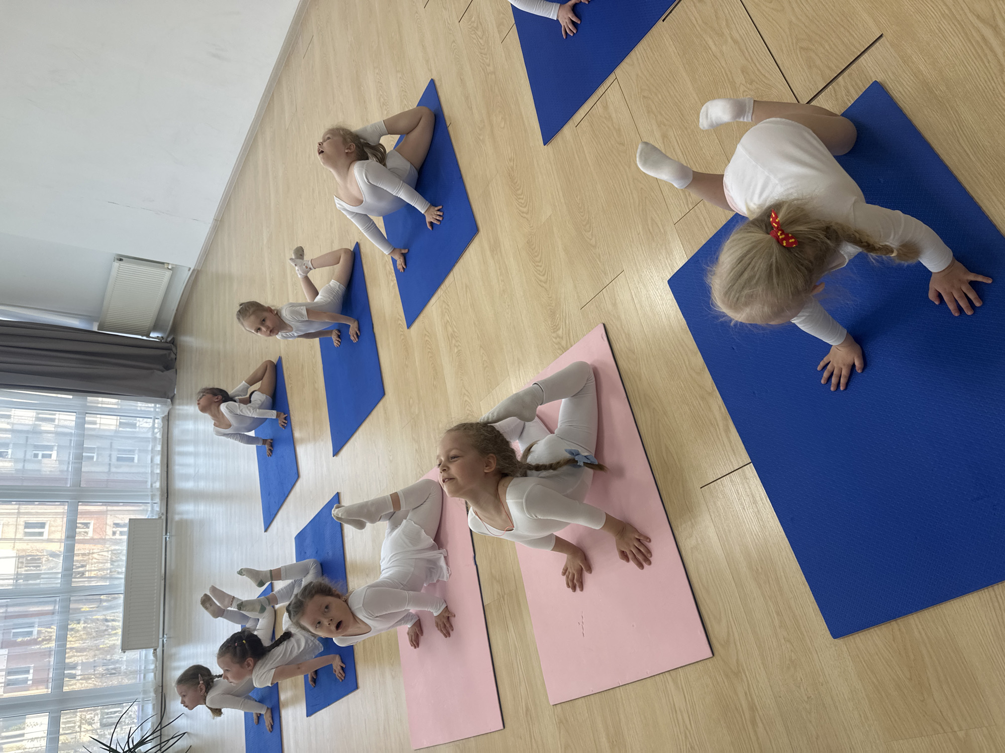 A group of young girls in white leotards are participating in a gymnastics or yoga class in what appears to be a dance studio or gymnasium in Votkinsk, Russia. The scene shows the girls positioned on individual mats, some blue and some pink, performing a specific pose that requires flexibility in the back and legs. Their faces show varying degrees of concentration and effort. The room has a light-colored wooden floor and natural light streaming in from a large window, suggesting a daytime setting. The studio's setting within Votkinsk is inferred, but not explicitly revealed by details in the image.