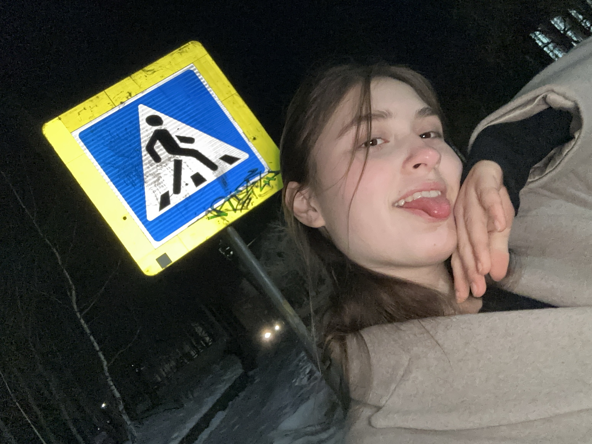 A young woman poses next to a pedestrian crossing sign in Sarapul, Russia at night. The sign is square, with a yellow border and a blue center featuring a white triangle containing a black pedestrian figure crossing white stripes. The woman, fair-skinned with brown hair, sticks her tongue out playfully. She wears a beige coat and a black top. The environment is dark, with bare trees visible in the background. Snow is visible at the bottom left of the frame. Two lights are visible in the distance.