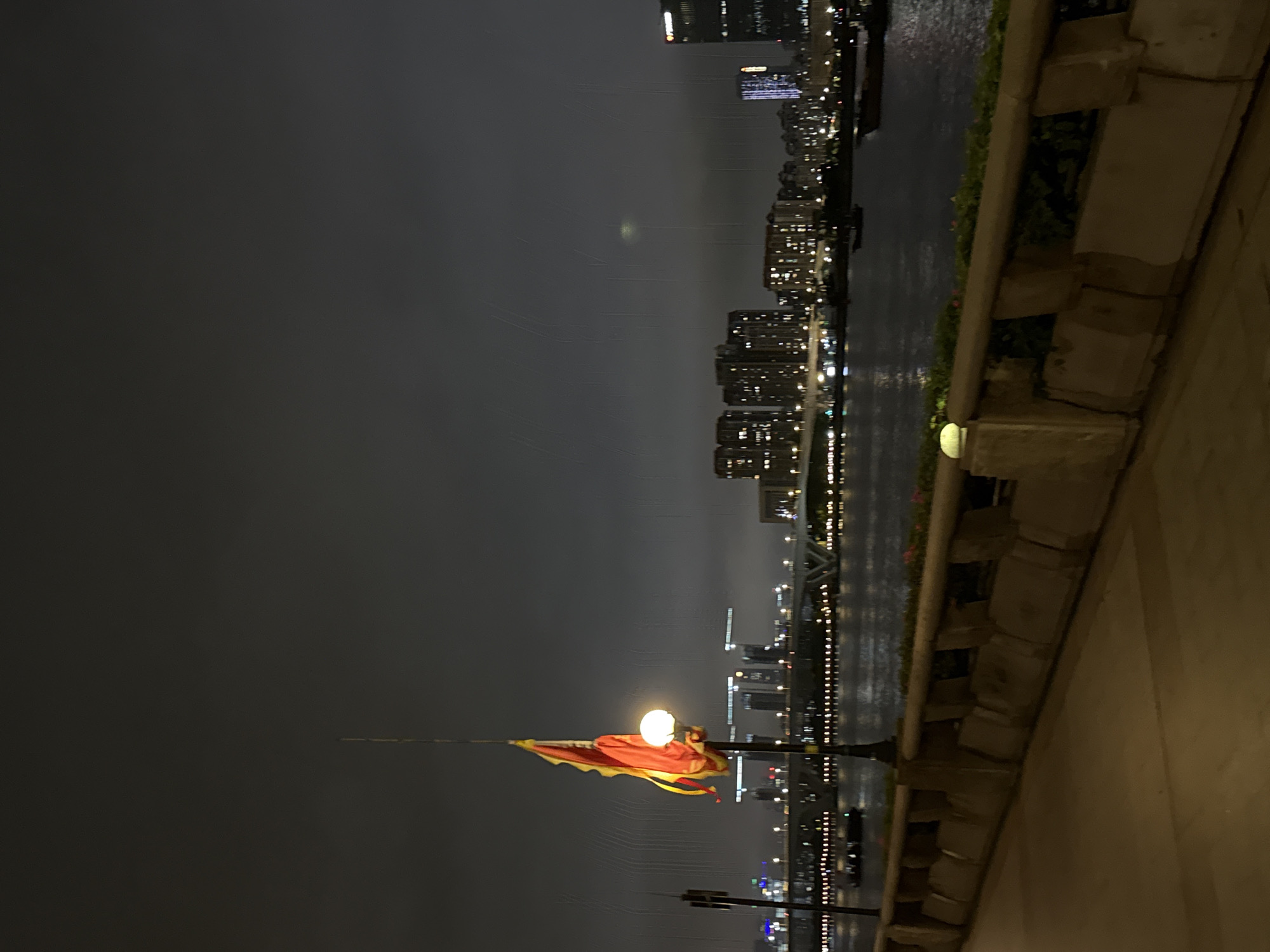 The image depicts a night scene in Haizhu District, China, with a cityscape overlooking a body of water. Tall buildings with illuminated windows form a prominent backdrop, suggesting an urban environment. The water's surface reflects the city lights, creating a shimmering effect.
In the foreground, a flag with red and yellow sections is visible, attached to a flagpole that also supports a bright light fixture. The flag appears to be gently waving. Beyond the flag, a bridge is illuminated with numerous lights, stretching across the water. The overall atmosphere is one of nocturnal urban activity.
The time of day is clearly night due to the artificial lighting and the dark sky. The presence of a slightly hazy or misty quality in the air, particularly in the upper portion of the image, might indicate atmospheric conditions or light diffusion. The foreground features a textured stone ledge and a concrete walkway, suggesting a pedestrian area or a waterfront promenade. No people are discernible in the image. The scene conveys a sense of calm and the vibrancy of a city at night. No visible text is present in the image.