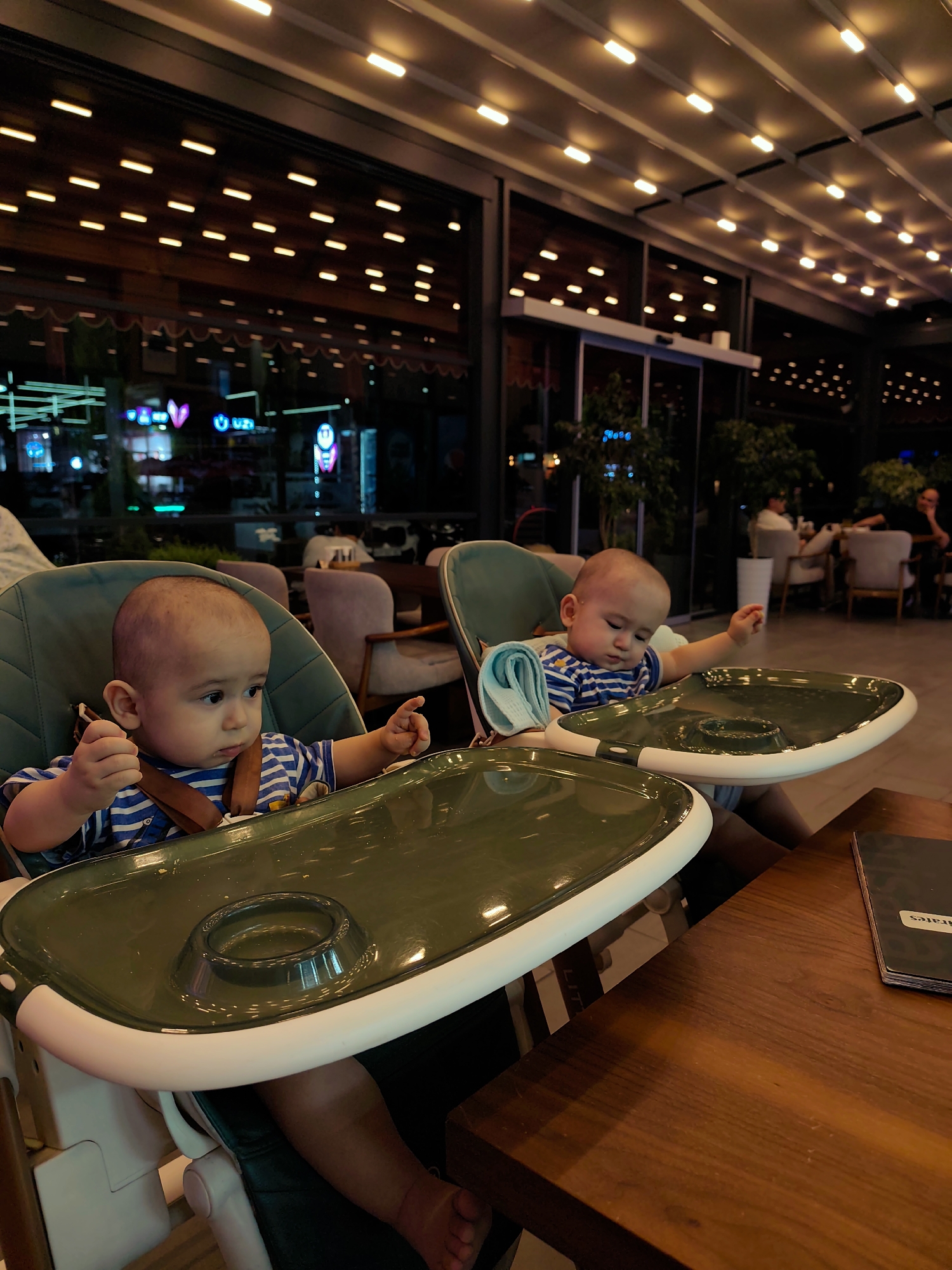 Two young children are seated in high chairs at a restaurant. The child on the left is looking to the right with a thoughtful expression, their hand resting on the high chair tray. The child on the right is looking down with their eyes closed, appearing sleepy, with a blue cloth draped over their shoulder. Both children are wearing striped shirts. The high chairs are green with white trim and have built-in trays. In the background, the restaurant appears to be open-air, with a retractable roof illuminated by rows of warm-toned lights. Through the glass facade, neon signs and lights from other establishments are visible, suggesting an urban setting at night. The overall atmosphere is dimly lit and cozy, with other patrons seated at tables in the background. A wooden table is in the foreground, and a menu with the word "pirates" visible on it is on the table. The time of day is evening.