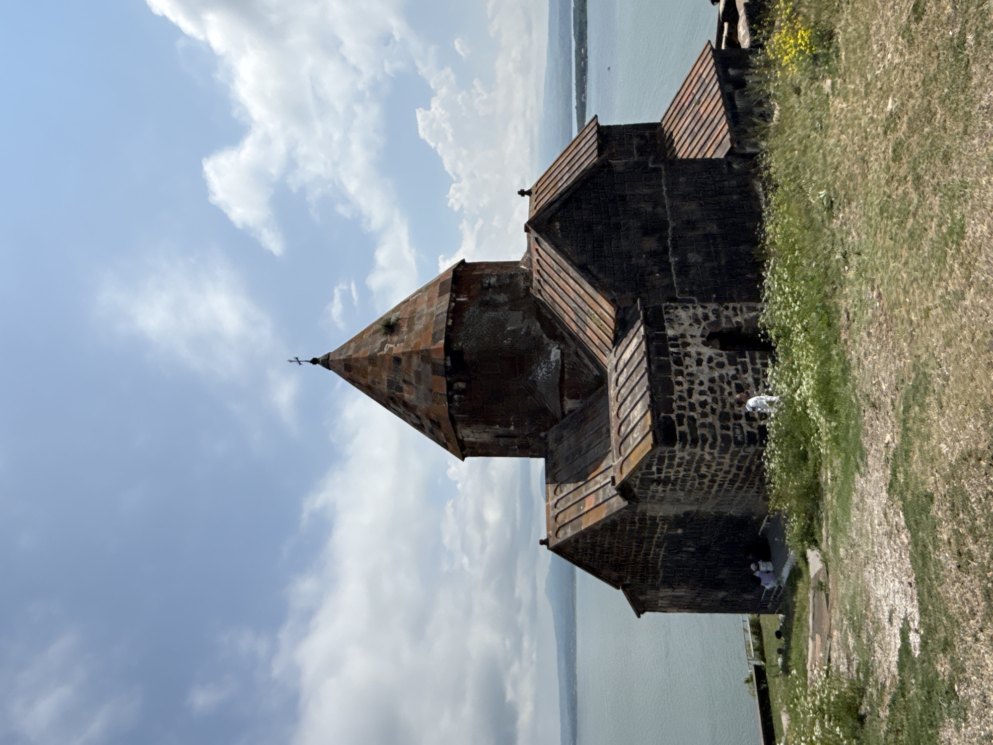 The image shows a stone church with a conical roof, likely an ancient structure, situated on a grassy hillside overlooking a body of water. The architecture is dark stone with a weathered, reddish-brown tiled roof. The sky is blue with scattered white clouds, suggesting a pleasant, possibly warm day. The scene is oriented vertically, with the water and a distant horizon line appearing at the top of the frame, and the ground, covered in dry grass and some wildflowers, filling the right side. Several small figures of people are visible near the base of the church, interacting with the structure or the surrounding area. One person appears to be leaning over a wall, while others are seen walking or standing. The overall impression is one of historical significance and natural beauty, with the church standing as a prominent landmark against the backdrop of the landscape. The time of day appears to be daytime, with the lighting casting some shadows but not indicating dusk or dawn. The location, identified as Sevan, Armenia, likely refers to a monastery or church near Lake Sevan, known for its dramatic setting. There is no visible text in the image.