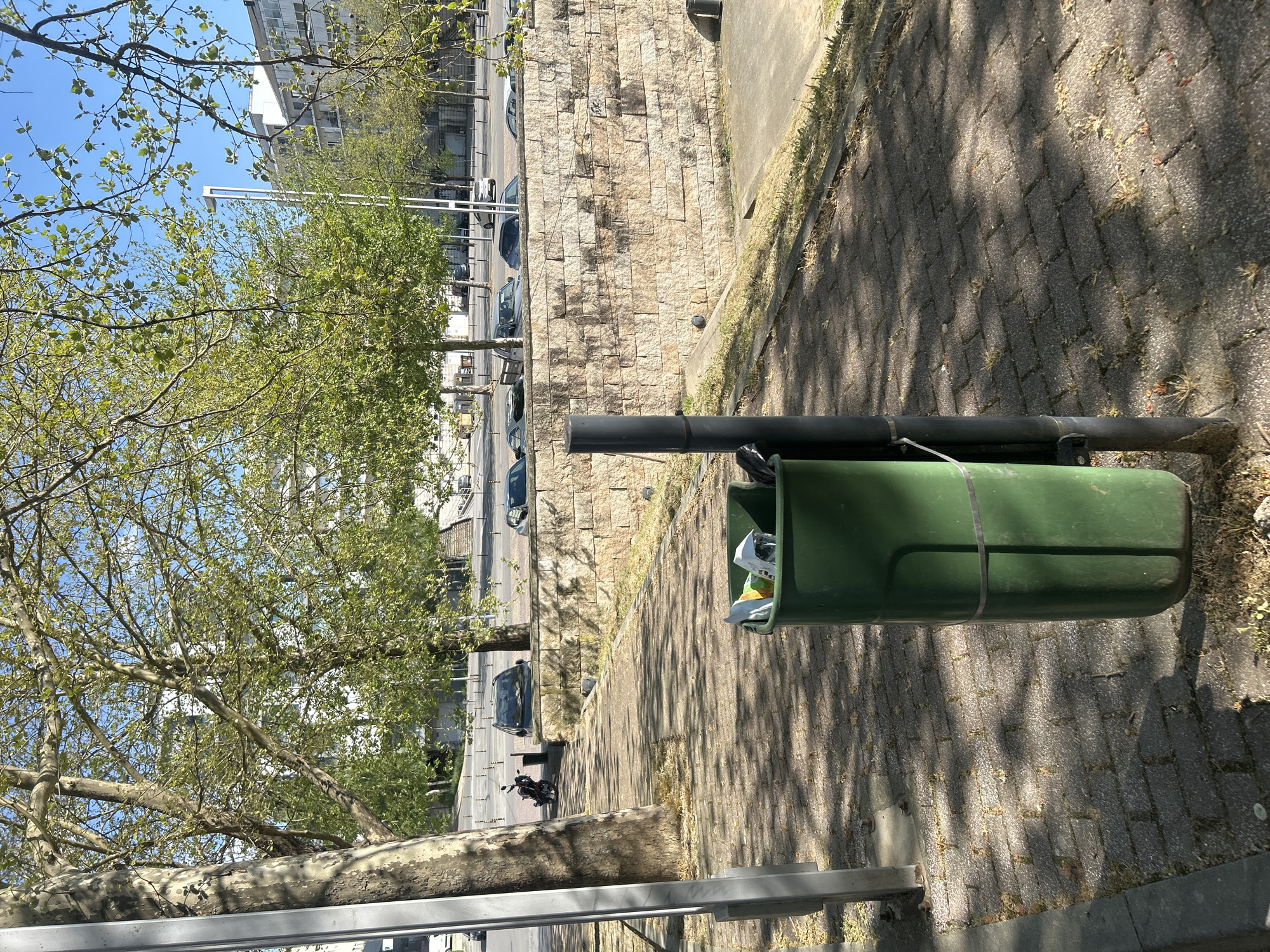 The image shows a street scene in Vila Real, Portugal. A green trash can is the prominent object in the foreground, secured to a metal post. The street behind the trash can is lined with trees with young leaves, casting shadows on the brick pavement. Several parked cars and a motorcycle are visible along the street. A stone wall rises above the street level. In the background, a tall building can be seen under a blue sky. The image is taken from an elevated perspective.