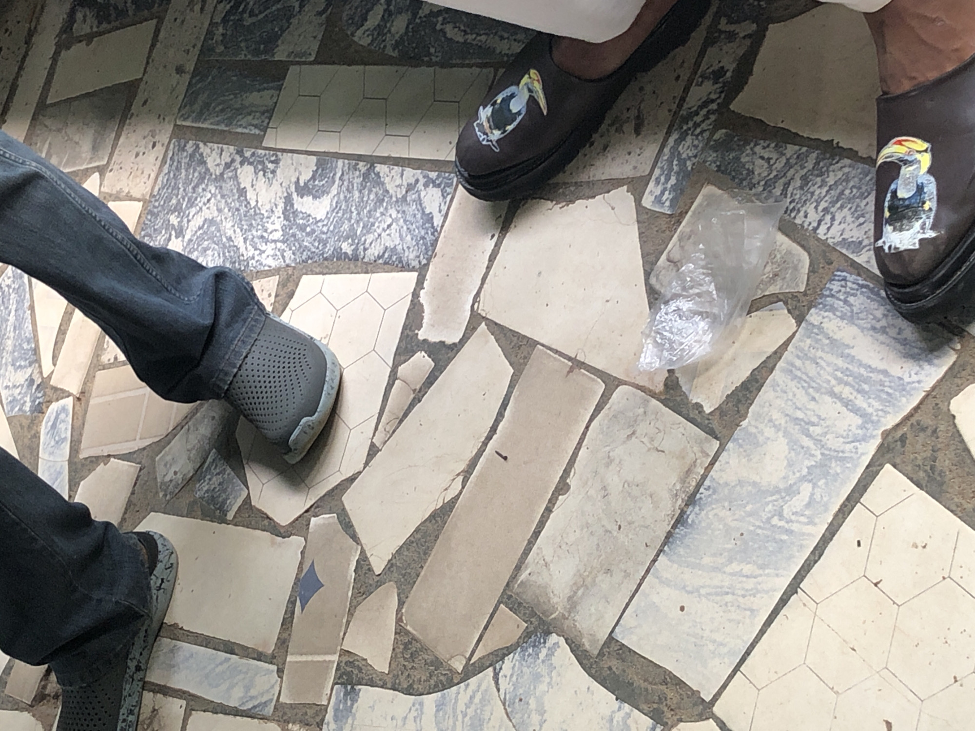 The image shows a close-up of a person's legs and feet standing on a floor made of irregular, mosaic-like tiles in various shades of cream, beige, and blue-gray with dark grout. The person is wearing dark denim jeans and two different types of footwear. On the left, the person is wearing a dark gray perforated shoe. On the right, the person is wearing a brown slip-on shoe with a decorative sticker of a yellow and blue cartoon character on it. There is a crumpled piece of clear plastic on the floor between the person's feet.