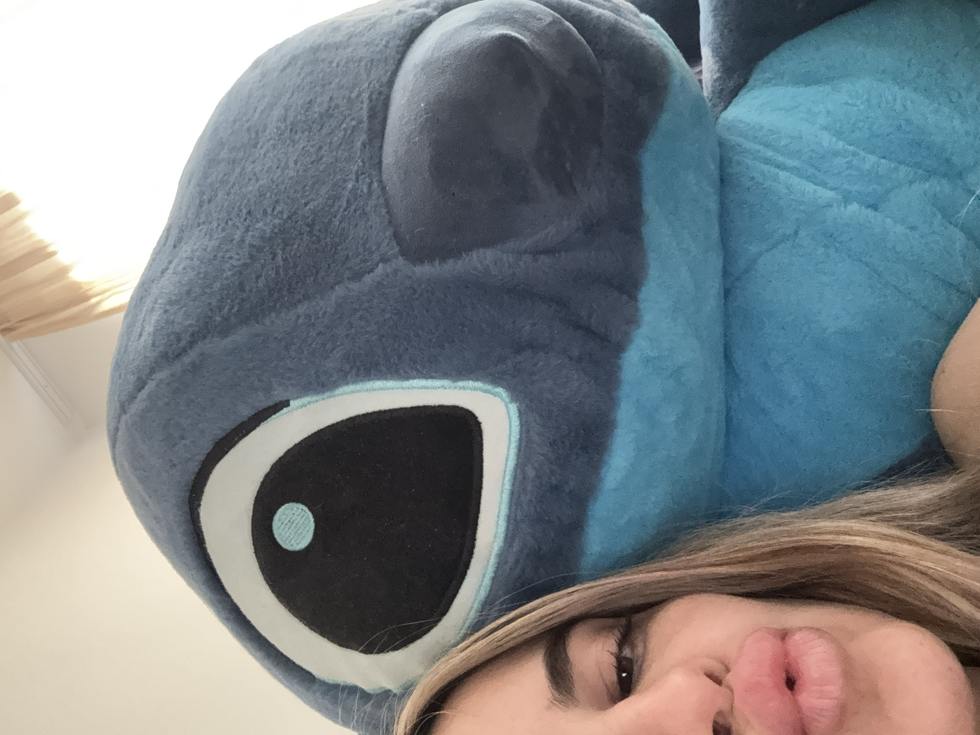An upward-facing selfie captures a person alongside a large, plush toy. The prominent subject is a soft, blue plush toy, resembling the character Stitch, which occupies the upper and left two-thirds of the frame. Its large black eye with a white and light blue iris and a light blue pupil is clearly visible, oriented towards the camera. A smaller, darker blue ear of the toy is visible above its eye.
In the bottom right corner, a portion of a person's face is visible. This includes their right eye, a well-defined eyebrow, part of their nose, and puckered lips, indicative of a "duck face" selfie pose. Light brown hair frames the person's face and extends over parts of the plush toy.
The background is predominantly a bright, light-colored surface, likely a wall or ceiling. In the top-left corner, a blurred, light-brown, textured object, possibly part of a window blind or light fixture, suggests an indoor setting. The bright ambient light indicates it is daytime. The activity is a casual selfie, likely taken while lying down. No visible text or specific cues for Sofia, Bulgaria are present in the image. The overall mood is playful and intimate.