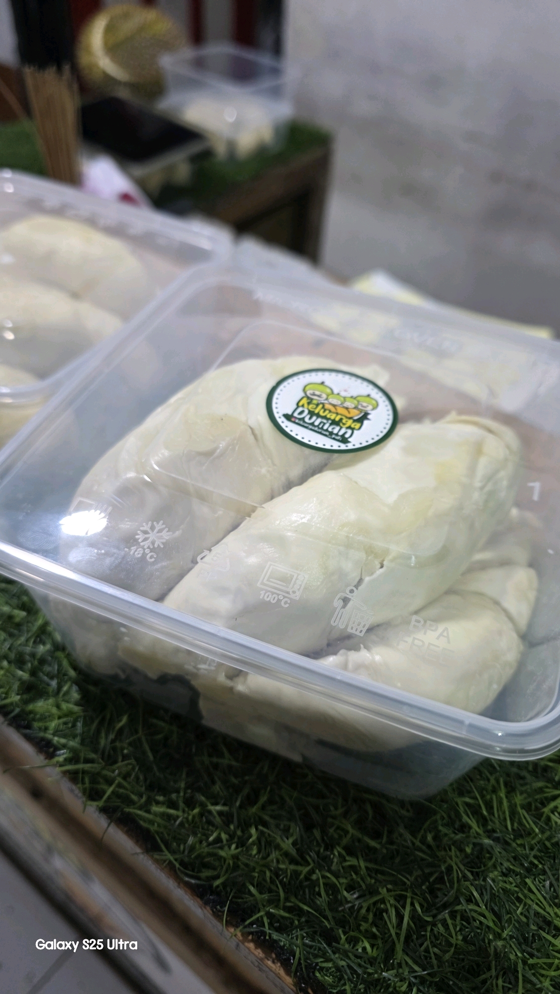In Kecamatan Pati, Indonesia, a close-up shot captures several durian flesh segments neatly arranged within a clear plastic container. The container has markings indicating it can withstand temperatures up to 100°C and is BPA-free. A circular sticker with a green border, depicting three cartoon durians and the text "Keluarga Durian," is affixed to the lid. The container rests on a surface covered with artificial green grass. In the blurred background, a whole durian and other plastic containers are visible. The bottom left corner displays "Galaxy S25 Ultra."