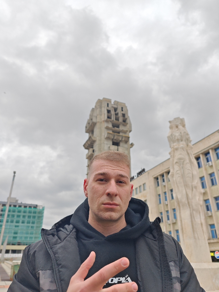 A male individual is depicted in the foreground, centered, facing the camera with a neutral expression. He has a very short, light-colored haircut and is wearing a black hooded sweatshirt beneath a black puffer jacket with camouflage detailing on the shoulders. His right hand is raised, forming a gesture with the thumb, index, and middle fingers extended.
The background prominently features the Monument to 1300 Years of Bulgaria in Shumen, Bulgaria, appearing as a large, brutalist concrete structure with geometric forms. To the right of this monument, a tall, white, abstract or figural sculpted monument is partially visible. Behind these monuments, a multi-story building with numerous windows and a light-colored facade is present; a partial "Pepsi" logo is visible on its lower section. To the far left, a building under construction or renovation, covered in green netting, is visible, with a long, slender pole extending upwards near it. The sky is overcast with grey clouds, indicating a cloudy outdoor environment.
