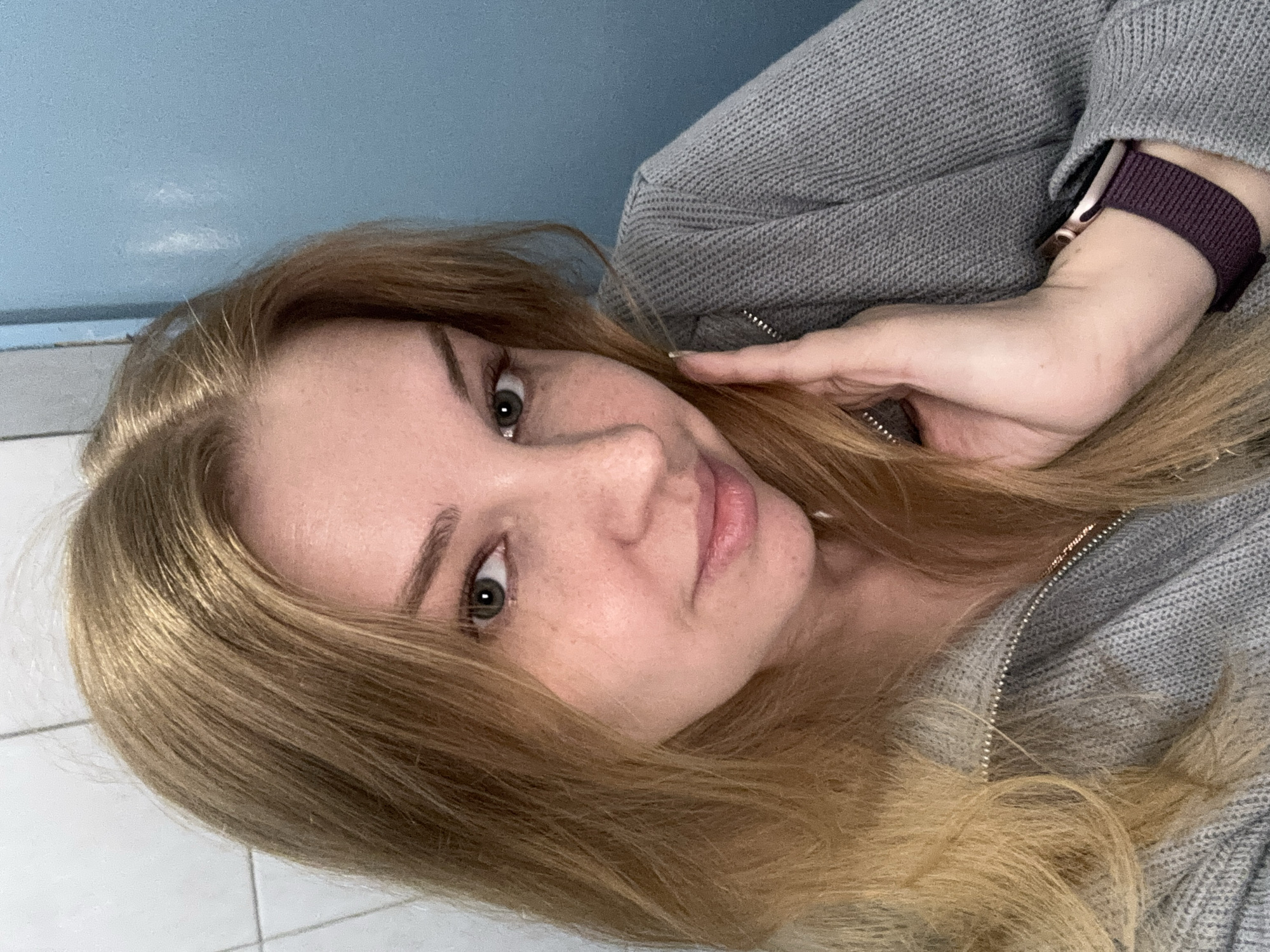A woman with long, light-brown hair is posing for a selfie. She is wearing a gray, knitted sweater and an Apple Watch with a purple band. The background is blue on the top and white tiles on the bottom, suggesting a bathroom.