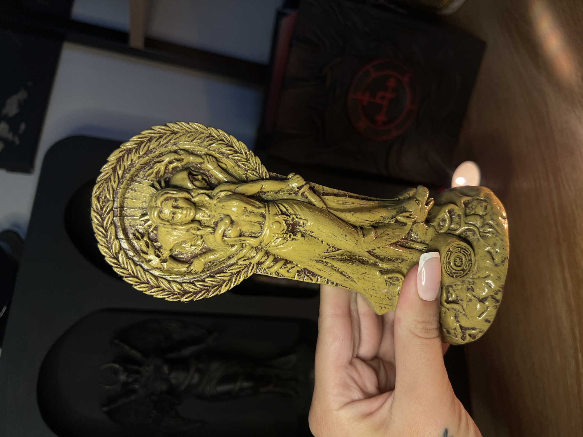 A hand is holding a golden statue of a multi-armed goddess wearing a crown and laurel wreath. The goddess has a snake wrapped around her torso. The statue is on a pedestal with intricate carvings. In the background, there is a dark silhouette of a winged figure with horns, and a black book with a red circular symbol. A small white candle is lit in front of the book. The scene appears to be on a wooden surface, likely a shelf. The location is Unknown, Unknown.