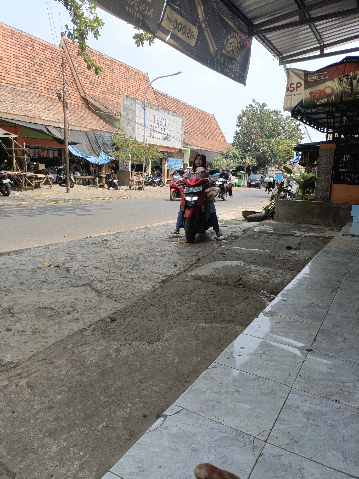 The image depicts a street scene in Betek, Indonesia. A woman and a child are seated on a red motorcycle stopped on the side of the road, near a market area with awnings and stalls. The market building has a tiled roof and a sign that reads "PASAR CONDONG PROBOLINGGO". Various motorcycles are parked in front of the stalls, and other vehicles are visible on the road. The ground in the foreground is paved with concrete, showing cracks and some debris. In the background, trees and buildings are visible under a bright sky. The overall impression is a bustling marketplace in a tropical environment.
