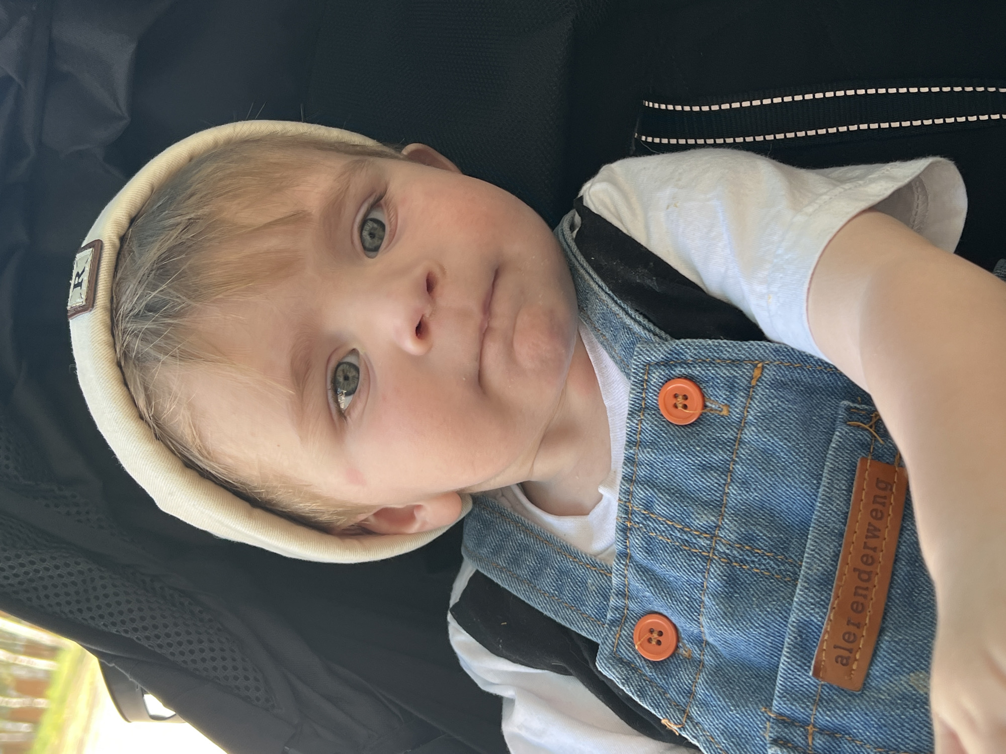 A close-up, slightly canted, medium-shot photograph depicts a toddler lying in what appears to be a stroller or similar conveyance. The child is fair-skinned with light-colored hair, wearing a cream-colored beanie, a white t-shirt, and denim overalls with orange buttons. The overalls have a brown leather tag with the word "alerenderweng" stitched on it. The backdrop is a dark, possibly black, material. The environment is partially visible in the lower left corner and suggests an outdoor setting with greenery. The toddler's expression is thoughtful, and their gaze is directed upwards. The location cannot be determined as Noviy, Russia, based on the image alone.