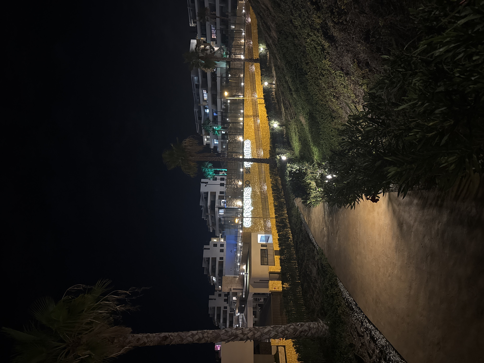 The image captures a nighttime view of the "Camposol Serentino" complex in Finestrat, Spain. A paved pathway, illuminated by soft ground lights, curves through lush green foliage on the right. To the left, a tall palm tree stands prominently against a dark sky. In the background, modern white buildings with multiple balconies are brightly lit. A yellow-lit wall with the complex's name, "camposol serentino," is visible, separating the foreground from the residential structures. The scene is serene and well-maintained, suggesting a resort or residential area.