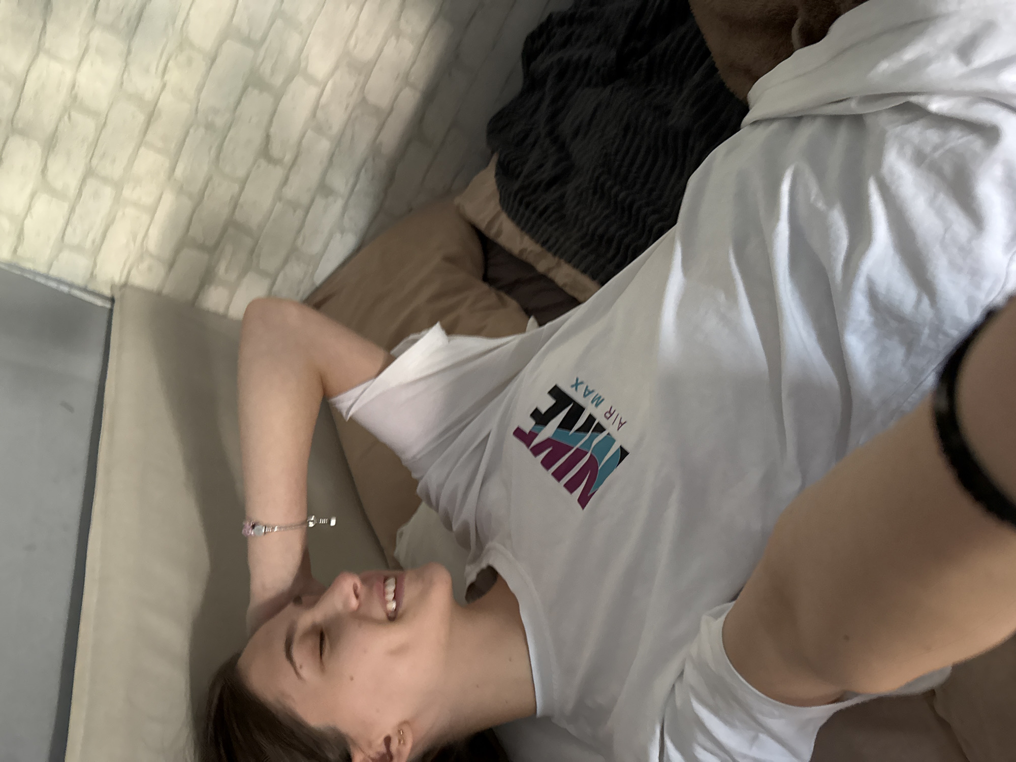 A young woman lies on a light gray couch, her head resting on it with her left arm raised. She wears a white t-shirt with a Nike Air Max logo. The background includes a brick-patterned wall and a dark gray blanket. The location is presumed to be indoors, potentially a residence in Sofia, Bulgaria, based on available information. The woman is smiling and appears relaxed.