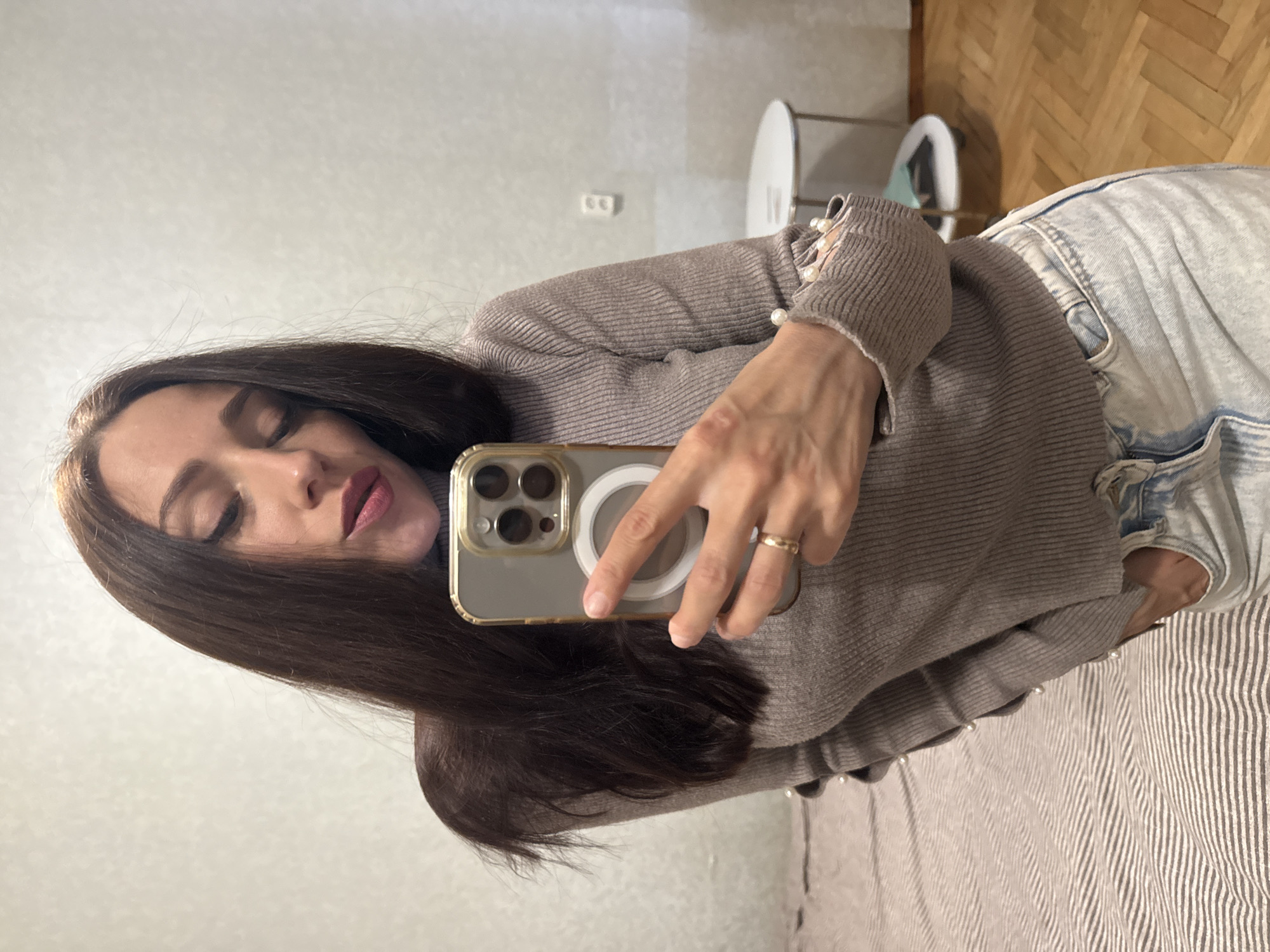A young woman with long, dark hair and light makeup, wearing a taupe turtleneck sweater with pearl embellishments on the sleeves and light-wash jeans, is taking a selfie in what appears to be a bedroom. The room has light-colored walls, a striped bedspread, and a wooden floor. In the background, a small white table with a round top is visible. The woman is holding a smartphone with a prominent camera array and a white circular accessory. She is wearing a gold ring on her left hand. The overall lighting suggests an indoor setting, possibly during the daytime.