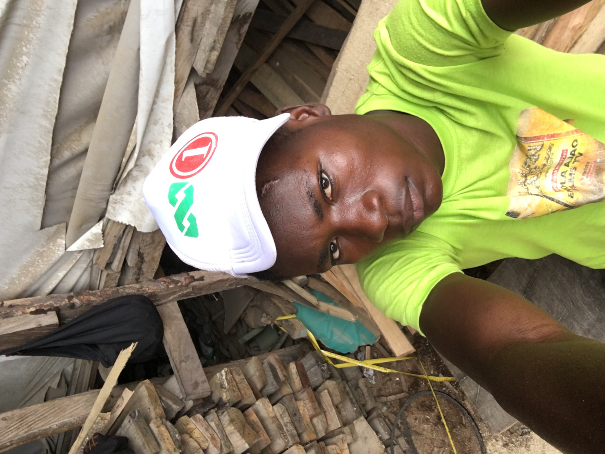 A close-up, upward-angled selfie captures a dark-skinned individual, likely male, in Ado, Nigeria. The person is positioned towards the right side of the frame, looking directly at the camera with a neutral to slightly pensive expression. A small, healed scar is visible on their forehead. They are wearing a bright neon green t-shirt and a white baseball cap.
The white cap features a distinctive logo: a red circle containing a white numeral "1", with green stylized letters resembling "GA" or "ZA" below it, possibly representing a political or organizational affiliation. Tucked into the front of the t-shirt is a crumpled piece of paper or label, clearly displaying the text "ENOLA AJAO TV" and "YouTube".
The background is a cluttered, rustic environment, suggestive of an outdoor or semi-sheltered area such as a construction site, lumberyard, or storage space. It is dominated by an array of rough-hewn wooden planks and pieces, stacked and strewn about. Light-colored, possibly soiled, fabric is draped and piled among the wood. Other visible objects include a black, folded bag-like item, a yellow measuring tape extending across the ground, and what appears to be a black cable or hose. The bright, even lighting indicates it is daytime.