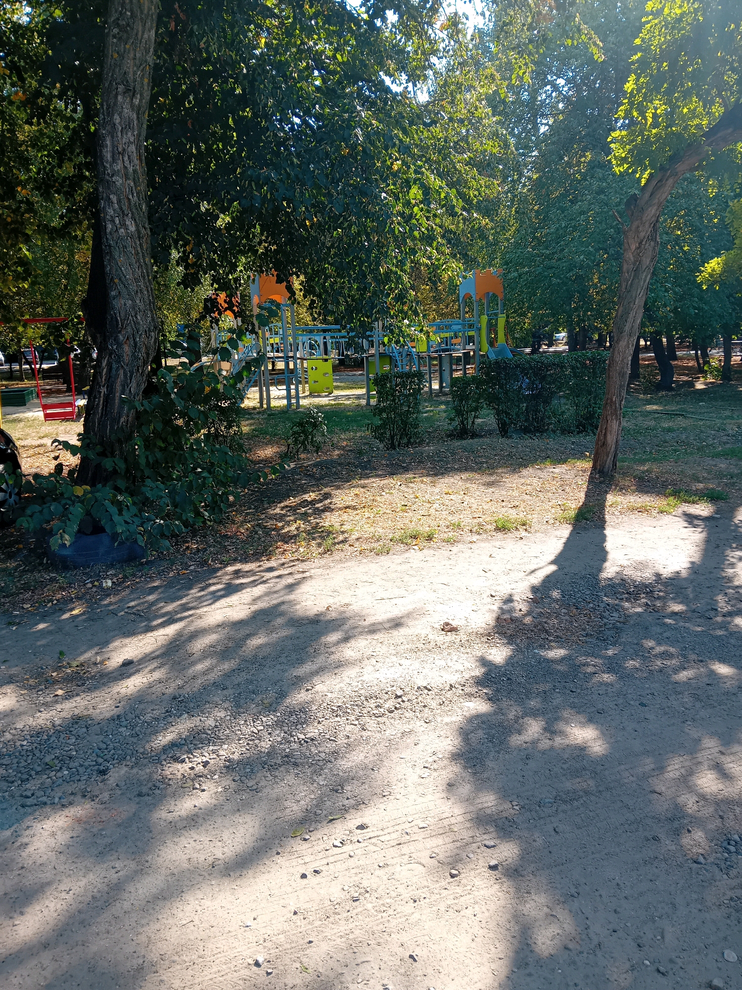 The image shows a playground area within a park setting, likely in Chuhuiv, Ukraine, based on the provided context. In the foreground, a dirt and gravel path is visible, dappled with shadows cast by the surrounding trees. To the left, the base of a large tree is prominent, with lush green bushes growing around it. Further into the scene, a playground structure with colorful elements like yellow, blue, and orange slides and climbing frames can be seen. The playground appears to be situated on a grassy area, with more trees forming a canopy overhead, suggesting a warm, sunny day. The overall environment is natural and recreational.