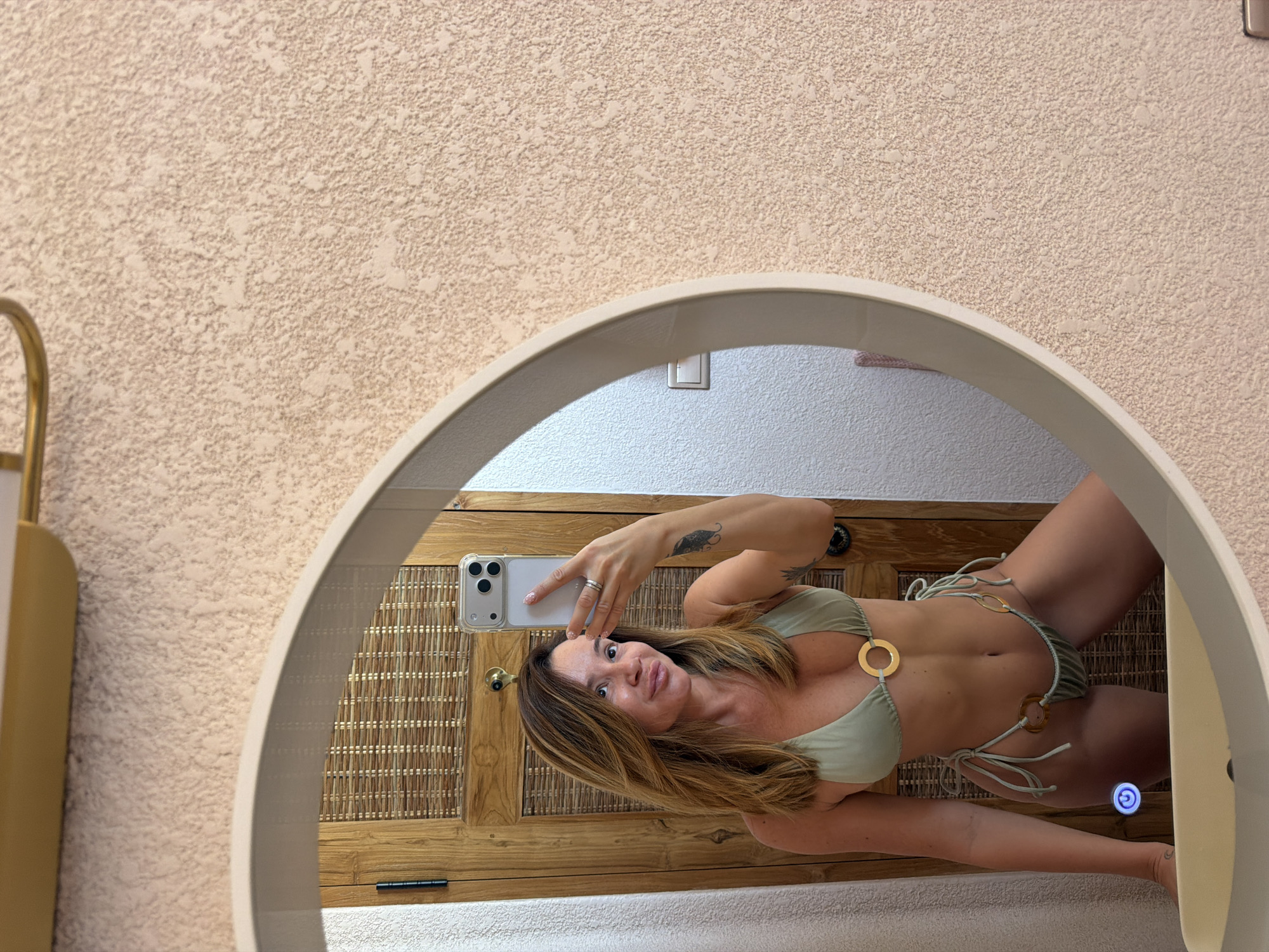 The image, rotated 90 degrees counter-clockwise, captures a woman taking a mirror selfie in what appears to be an indoor setting, likely a hotel bathroom or changing area in Mengwi, Indonesia.
The central subject is a woman with long, light brown hair, looking directly at her smartphone as she holds it up to take the photo. She wears a light green two-piece bikini featuring prominent gold circular O-rings connecting the top and bottom pieces. A ring is visible on her left-hand finger, and a dark tattoo of what appears to be a feather or abstract design is on her right upper arm. She has a slight smile on her face.
The mirror is circular or oval-shaped with a light-colored frame. Reflected in the mirror behind the woman is a wooden cabinet or door with a woven rattan panel, suggesting a tropical or resort aesthetic. A light switch is visible on the wall above the cabinet, and a blue illuminated power-like symbol, likely a touch sensor for a mirror light, is reflected below her waist. The surface just below the mirror reflection appears to be a light-colored, possibly textured, towel or rug.
Outside the mirror's frame, the surrounding wall has a textured, light beige finish. A brass-colored fixture, possibly a light or part of a vanity, is partially visible on the left side of the frame. No discernible text is visible in the image.