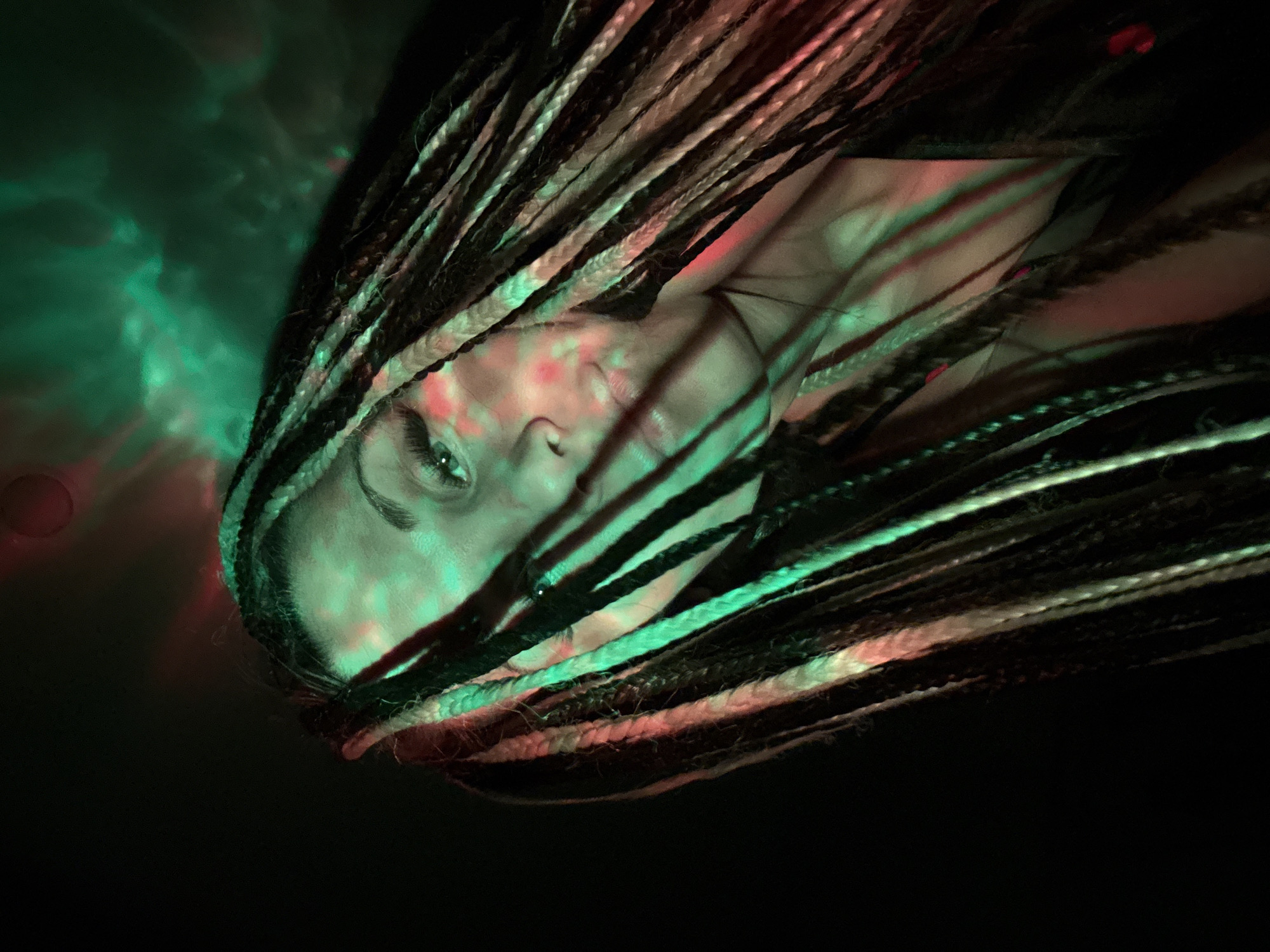 The image features a close-up portrait of a person, likely a woman, with her face partially visible and framed by numerous dreadlocks or braids. Her left eye is open and discernible, gazing towards the upper left of the frame, with a calm or contemplative expression.
The most striking element of the scene is the dramatic, artificial lighting. Vibrant green and red hues are projected onto the subject's face, hair, and the dark background. The lighting creates abstract, swirling patterns resembling clouds, smoke, or light refracted through water, particularly prominent in the upper left. Specific spots of red light are visible on her cheek and forehead. The dreadlocks themselves show variations in color, including dark, lighter, and reddish tones, which are further emphasized by the colored illumination.
The setting appears to be a low-light or dark environment, suggesting night, an indoor space, or a specialized studio setup. The dynamic and colorful lighting implies a context such as a club, concert, artistic performance, or a themed photoshoot. The image is composed with a strong diagonal tilt, adding to its artistic and dynamic feel.
No specific activity beyond being present and illuminated in this environment is depicted. There is no visible text. The location context provided is Rusanovo, Russia, although the image does not contain specific visual cues to confirm this geographical detail.