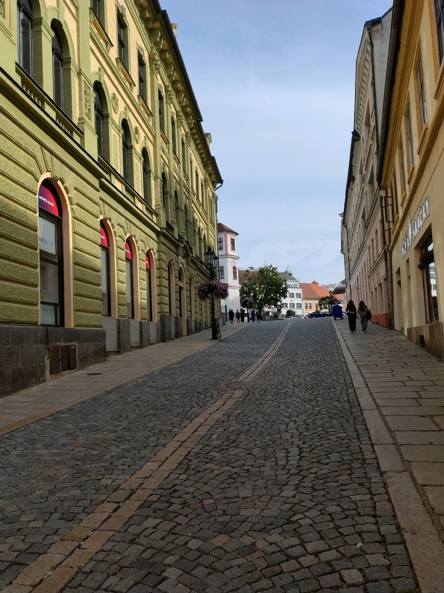 The image captures a cobblestone street in the historic center of Písek, Czechia, likely during daylight given the illumination and clear sky. The perspective is from street level, looking uphill. On the left, a light green building with arched windows and decorative detailing lines the street. A sign for "MONETA BANK" is visible in one of the windows. The building's facade exhibits textured plasterwork. On the right side, a yellow-toned building with similar arched windows and architectural embellishments also runs alongside the street. The street itself is paved with cobblestones, with a distinct yellow line of contrasting cobblestones marking a boundary. Further up the street, several people are walking. In the distance, other buildings, trees, and a blue trash bin are visible. A lamppost with hanging flowers stands near the green building. The scene depicts a typical European urban environment with historical architecture.