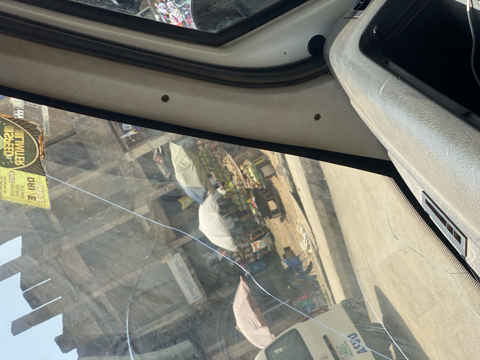 The image, taken from inside a vehicle, shows a street scene through a cracked windshield. In the foreground, the dashboard of the vehicle is visible, with part of the steering wheel and what appears to be a storage compartment. Outside the windshield, a bustling street market is depicted. Several stalls with umbrellas for shade are set up, displaying various goods, likely fruits and other produce. Some people are visible in the market, seemingly engaged in transactions or walking through the area. A white vehicle, possibly a van, with a license plate that includes "ASJO," is parked on the left side of the frame. In the background, unfinished structures and buildings suggest an urban environment. A yellow sticker with text in English, including "DRIVE SAFE" and "LIMITED," is affixed to the upper portion of the windshield. The overall impression is of a busy market day in Jos, Nigeria, viewed from the confined space of a vehicle.