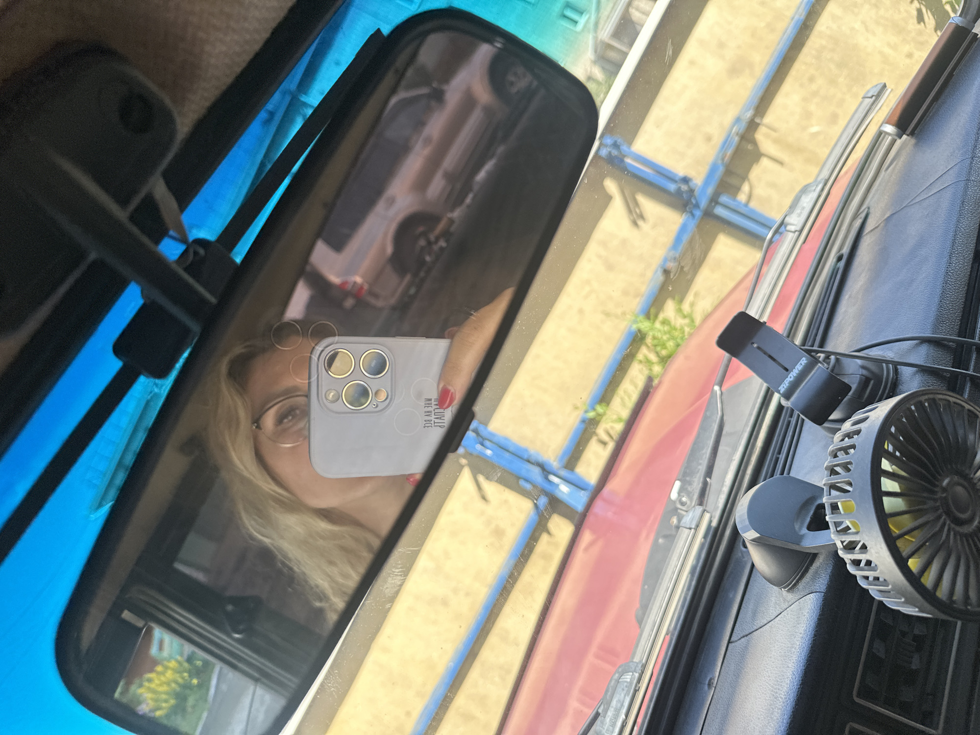 A woman is taking a selfie in the rearview mirror of a car. The car interior is visible, including a dashboard with a small fan and a phone mount. The woman has blonde hair and is wearing glasses. She is holding a phone with a light grey case and multiple camera lenses. Outside the car, a red vehicle and a tan SUV are visible, along with a blue fence and some greenery. The scene is likely in Novoaltaysk, Russia, given the context.