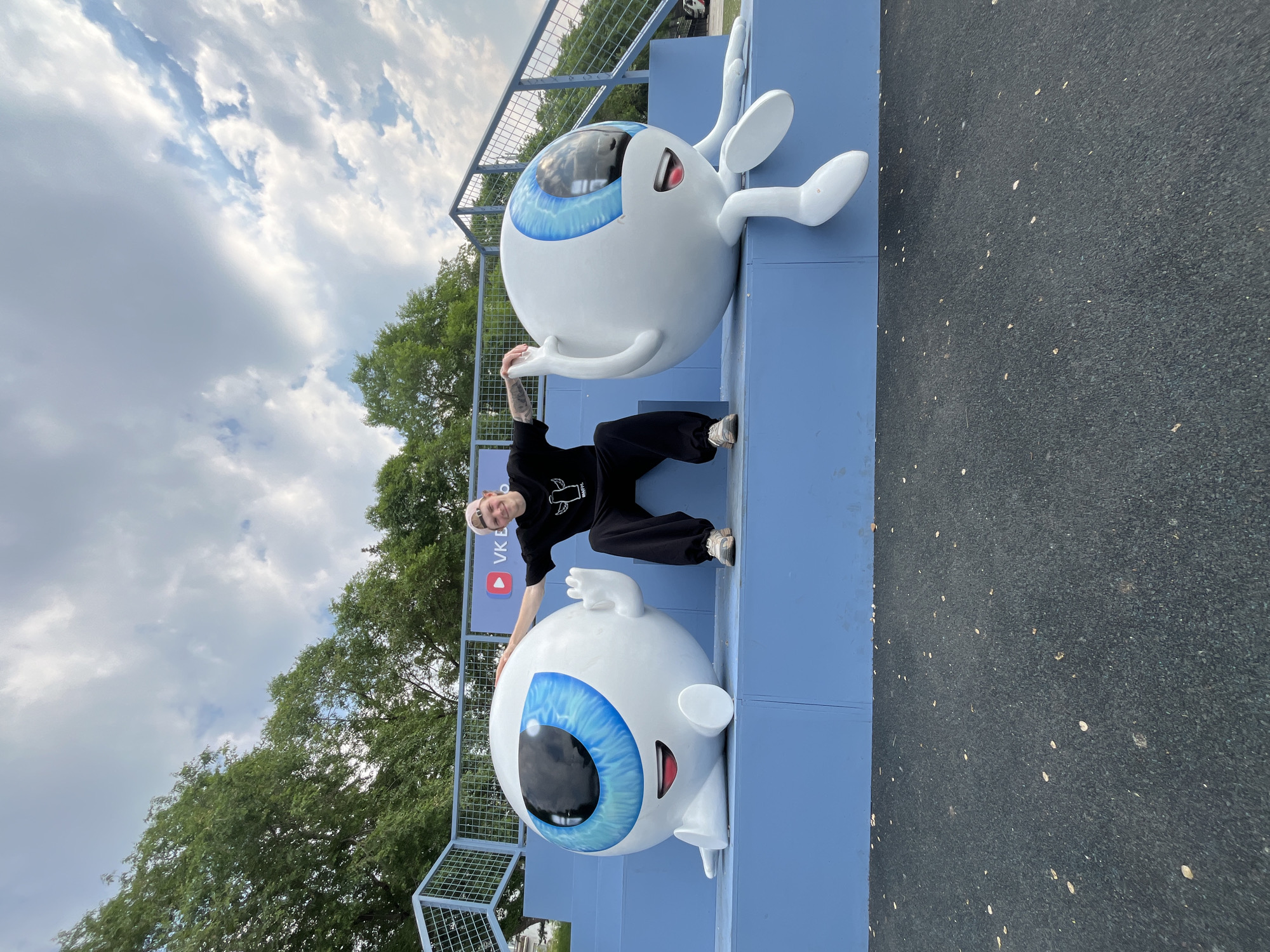 A smiling young person with blonde hair, wearing a black t-shirt and dark pants, sits on blue bleachers between two large, cartoonish eyeball statues. The statues have wide blue eyes with black pupils, open mouths with red lips, and white arms and legs. The person is positioned in the center, with one hand resting on the left statue's head and the other extended towards the right statue. Behind them, a fence and trees are visible, along with a cloudy sky. The ground in front of the bleachers is dark and textured, possibly a running track, with scattered small leaves. A banner with a VK logo is visible behind the statues. This scene appears to be at an outdoor event or park in Krasnoyarsk, Russia, possibly related to the VK social media platform.