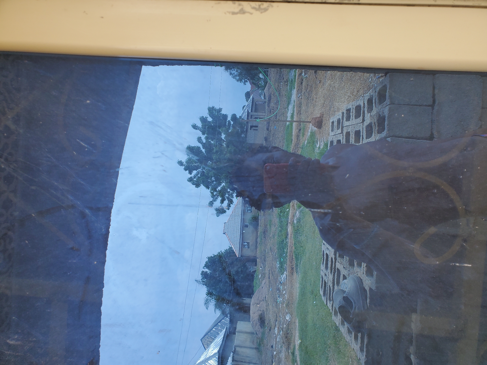 The image shows a reflection of a person holding a smartphone in their hand, presumably taking the photograph. The person's face is partially visible, and they appear to be shirtless. In the foreground, there is a pair of shoes placed on a stack of cinder blocks. The background of the reflection reveals a rural or semi-urban landscape with several buildings, some of which appear to be under construction or incomplete. Trees and vegetation are also visible, along with a cloudy sky. The scene suggests an outdoor setting, possibly in a developing area. The image does not contain any information that would allow for specific identification of the location within Winters, United States of America.