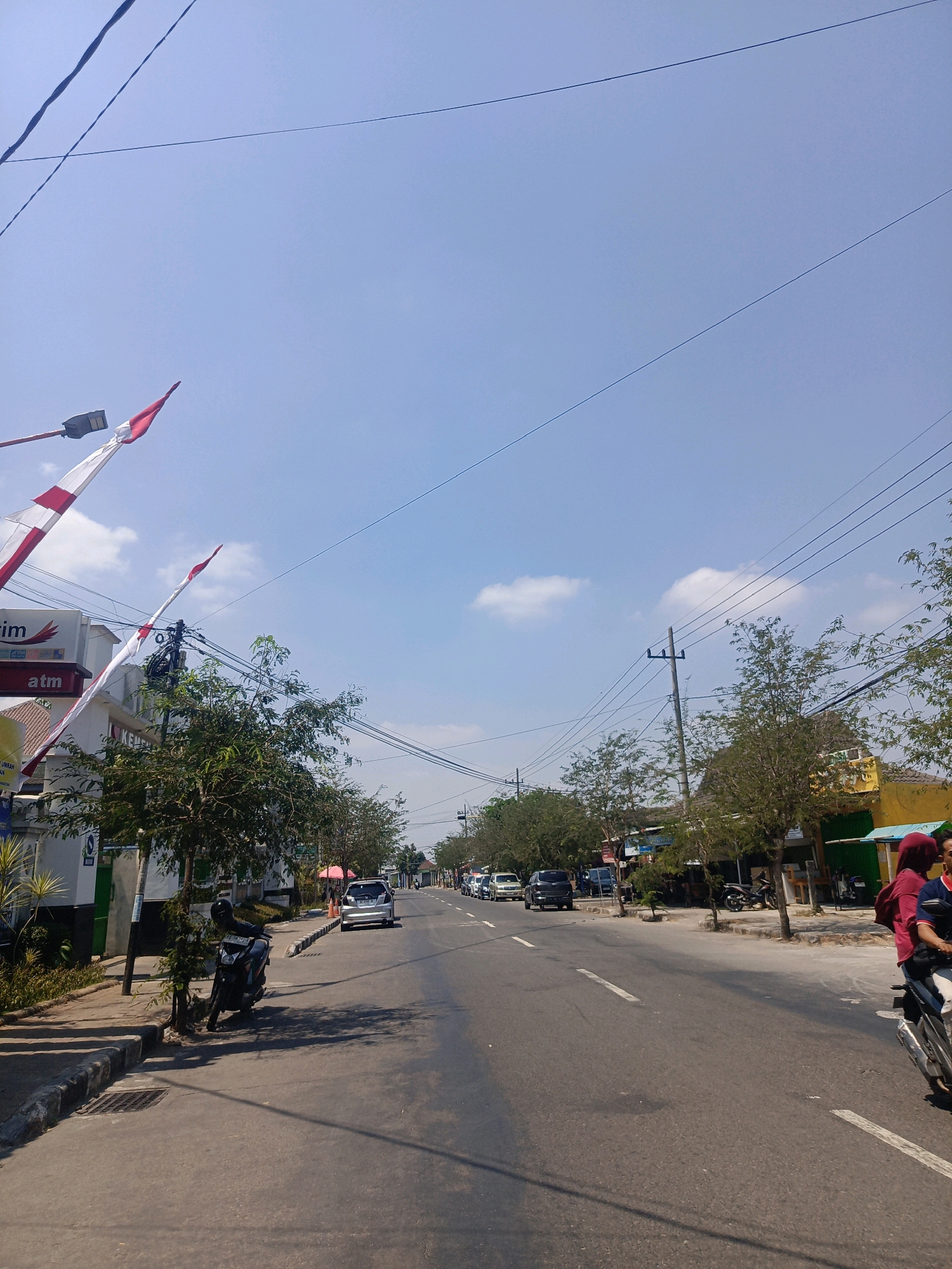 The image depicts a street scene in Sumberagung, Indonesia, on a bright, clear day. The road is paved and has white lane markings. On the left side of the road, there is a sidewalk with a low curb and a storm drain. A black motorcycle is parked on the sidewalk. Adjacent to the sidewalk are buildings, including one with a sign that says "ATM". Red and white flags are visible on poles above the buildings. Trees with green foliage line the sidewalk and extend along the street. On the right side of the road, more buildings are visible, some with awnings or open fronts. A few cars and motorcycles are present on the road, with traffic appearing to be moderate. Two people are riding on a motorcycle in the foreground on the right side of the image, with one person wearing a red headscarf and a red shirt. The sky is blue with a few scattered white clouds, and power lines are strung across the sky. The overall impression is a typical street view in a populated area of Indonesia.