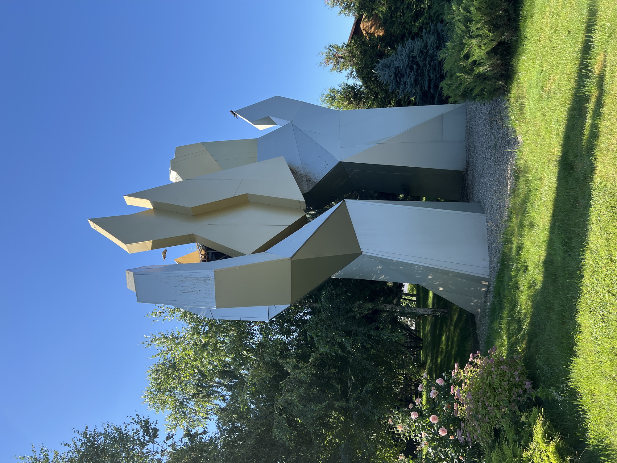 A large, abstract, metallic sculpture stands in a garden under a clear blue sky. The sculpture is composed of sharp, angular planes that are mostly silver with some beige accents. It has a dynamic, almost flame-like appearance, reaching upwards. The sculpture is situated in a well-maintained garden with lush green grass, manicured bushes, and a rose bush with pink flowers on the left side. Behind the sculpture, various trees, including birch and evergreen varieties, are visible, along with a hint of a wooden building to the far right. A small bird is perched on the top of the sculpture. The scene appears to be located outdoors, likely in a park or private garden setting. The image is taken from a medium distance, capturing the entire sculpture and its surrounding environment.