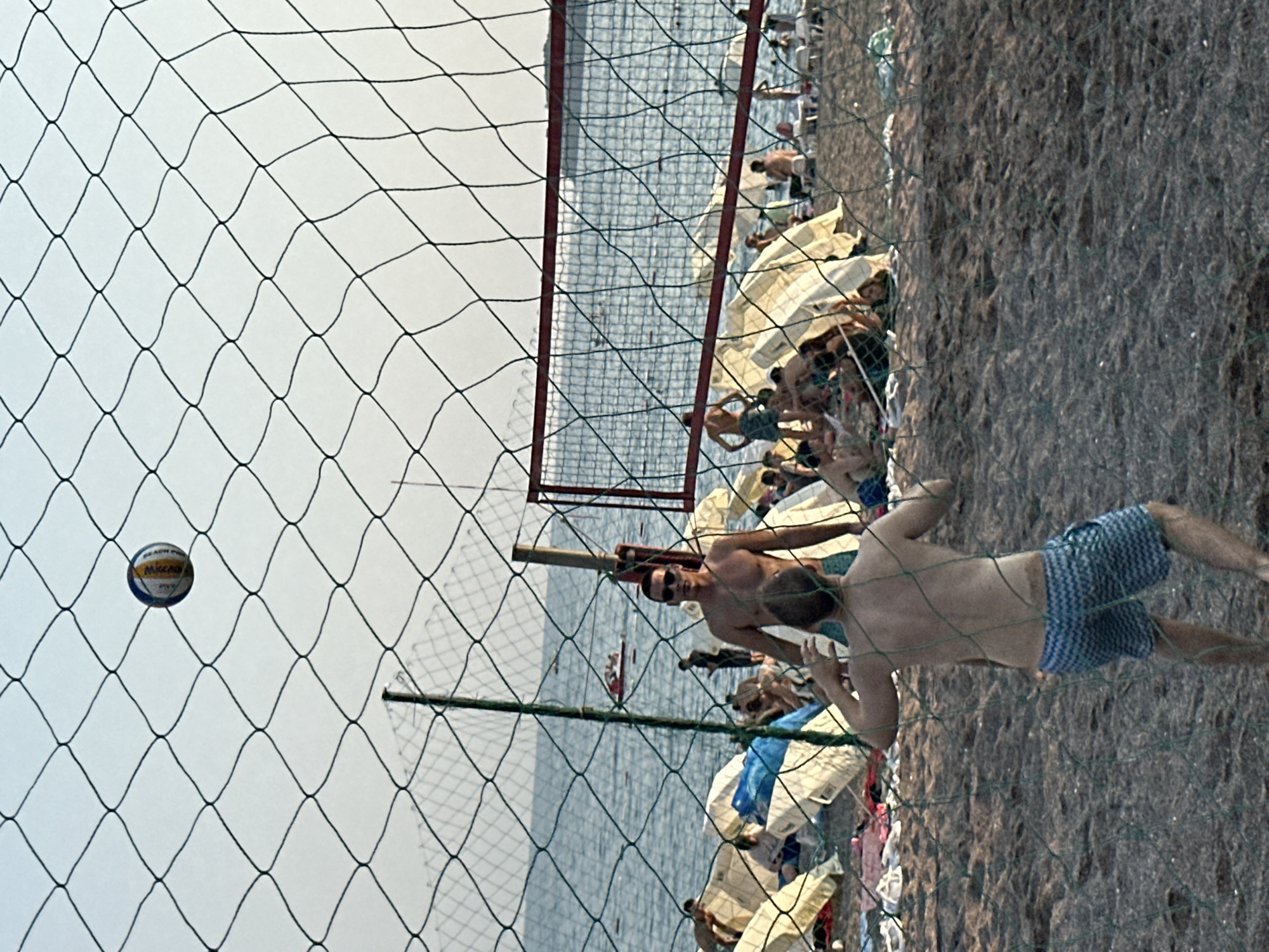 The image depicts a beach volleyball game at an outdoor location, likely in Boreti, Montenegro, given the context. The foreground is dominated by a green volleyball net. Beyond the net, two men are visible on the sandy court. One man, closer to the net, is in motion, with his back to the camera, seemingly preparing to receive a volleyball. He is wearing blue and white patterned shorts. The other man, further back and to the right, is standing, facing forward, wearing sunglasses and blue shorts. He is in a pose that suggests he has just served or is about to receive the ball.
In the background, the sea is visible with a few people and a small boat on the water. The beach is populated with numerous beach umbrellas and several groups of people relaxing on the sand. The overall atmosphere suggests a leisurely day at the beach. The lighting indicates it might be late afternoon or early evening.