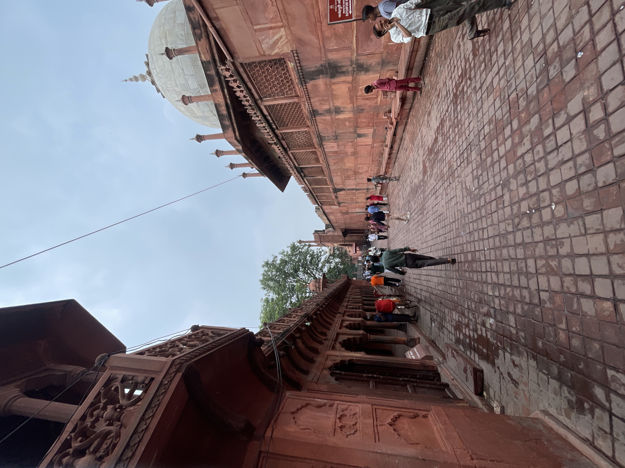 The image depicts a wide paved pathway lined with red sandstone buildings on either side, leading towards a large white dome. The pathway is wet, suggesting recent rain. Several people are walking along the path, some individually and some in small groups, dressed in casual clothing. The architecture is consistent with historical Indian structures, specifically resembling the Red Fort in Agra, India. One building on the left features intricate carvings and arched doorways. On the right, a section of the building shows latticed windows and is topped with decorative finials around the dome. A sign with Hindi text and an inscription "FATEHPURI MASJID" is visible on the right wall, confirming the location as Fatehpur Masjid within the Red Fort complex in Agra. The sky is overcast, with a light gray hue.