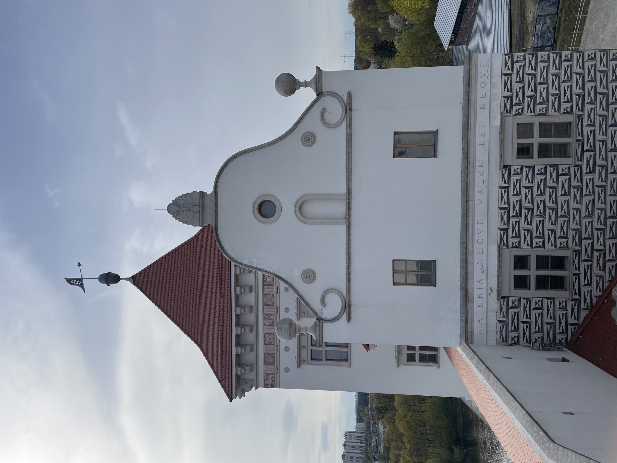 A historical building, likely a castle or palace in Hrodna, Belarus, is depicted from a slightly low angle against a bright, partly cloudy sky. The prominent structure features a stark white facade with a steep, red tiled roof. A decorative, curved gable crowns the top of the wall, adorned with a large spherical finial on the right and a shell-like ornament on the left. A black weathervane, bearing the year "1585," sits atop a spire on the main roof.
The white wall is embellished with intricate details, including scroll-like motifs, small rosettes, and bands of patterned relief above a series of small, corbel-like projections. Below the spherical finial on the right, a rectangular window with dark frames is visible. Another smaller, circular window and a recessed arched niche punctuate the upper part of the gable.
A horizontal band across the lower section of the white wall carries a Latin inscription in raised letters: "MATERIA S N E O V E MALVM EST N E O Q V E." Below this inscription, a significant portion of the wall to the right is covered in a bold, black and white geometric pattern, resembling sgraffito or painted decoration, framing a multi-pane window. Other windows with wooden frames are also visible on the white walls.
In the background, lush green trees indicate a spring or summer setting, and what appear to be distant urban structures and construction cranes can be seen. The lighting suggests daytime, possibly late morning or early afternoon, with soft, diffused sunlight. No human figures or discernible activity are present, emphasizing the architectural presence of the building.