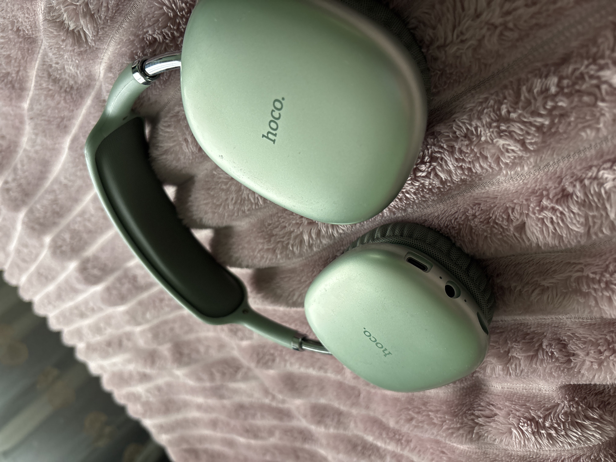 The image shows a pair of light green "hoco" branded over-ear headphones resting on a pink, textured blanket or throw. The headphones have silver metal arms connecting the earcups to the headband. The earcup cushions appear to be dark gray fabric. The location is not discernible from the image.