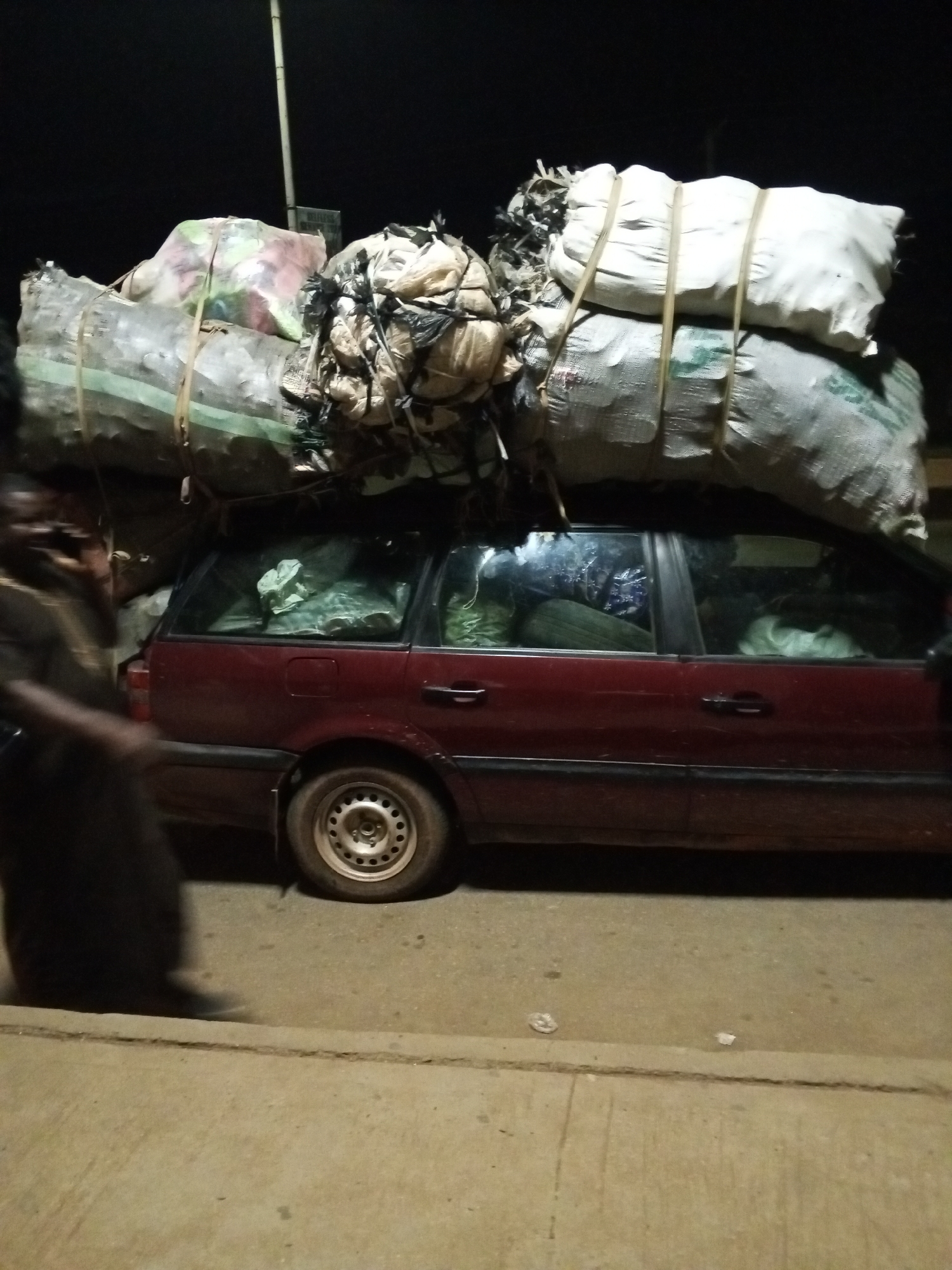 The photograph captures a dark maroon station wagon parked on a street in Bukuru, Nigeria. The car is overloaded with large sacks and bundled items secured to the roof, filling the interior as well. A person is walking past the car on the driver's side, only partially visible. The street is paved. A street light is visible in the background. The lighting suggests it is nighttime. The wheels of the car are visible, with standard hubcaps. The car's windows are tinted.