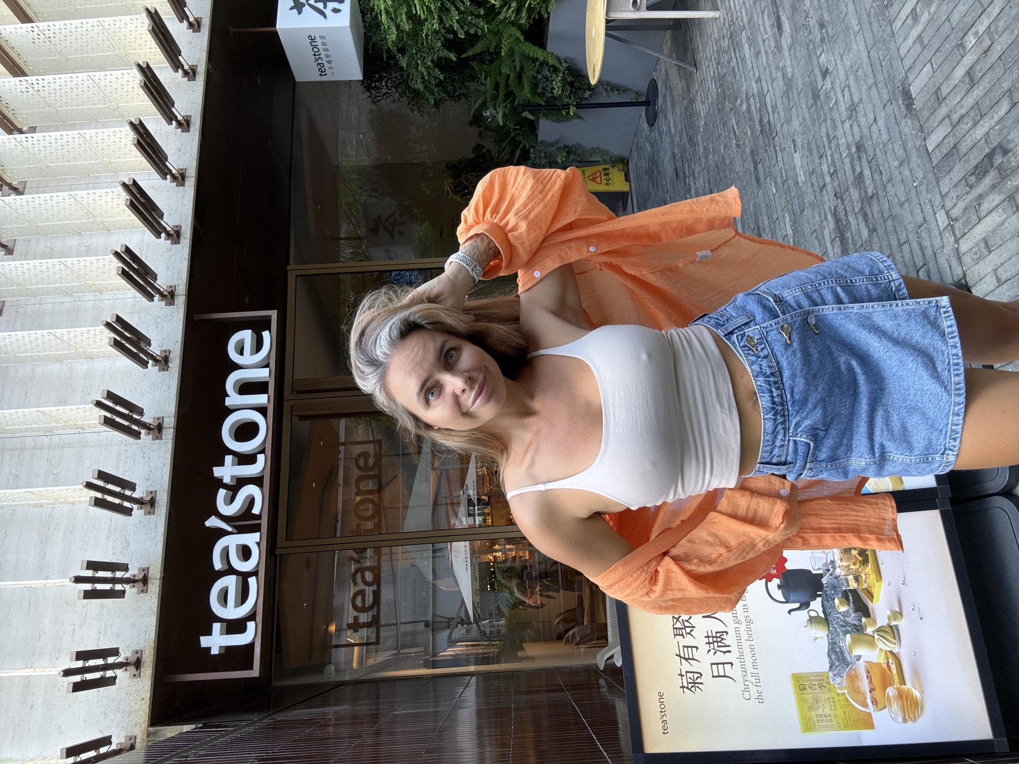 A woman with blonde hair streaked with gray is posing outside a building with a sign that reads "tea'stone." She is wearing a white tank top, an orange open button-down shirt, and a denim skirt. The building appears to be a tea shop, with a large poster displaying tea sets and Chinese characters. The background includes greenery and outdoor seating, suggesting a commercial area in Shanghai, China.