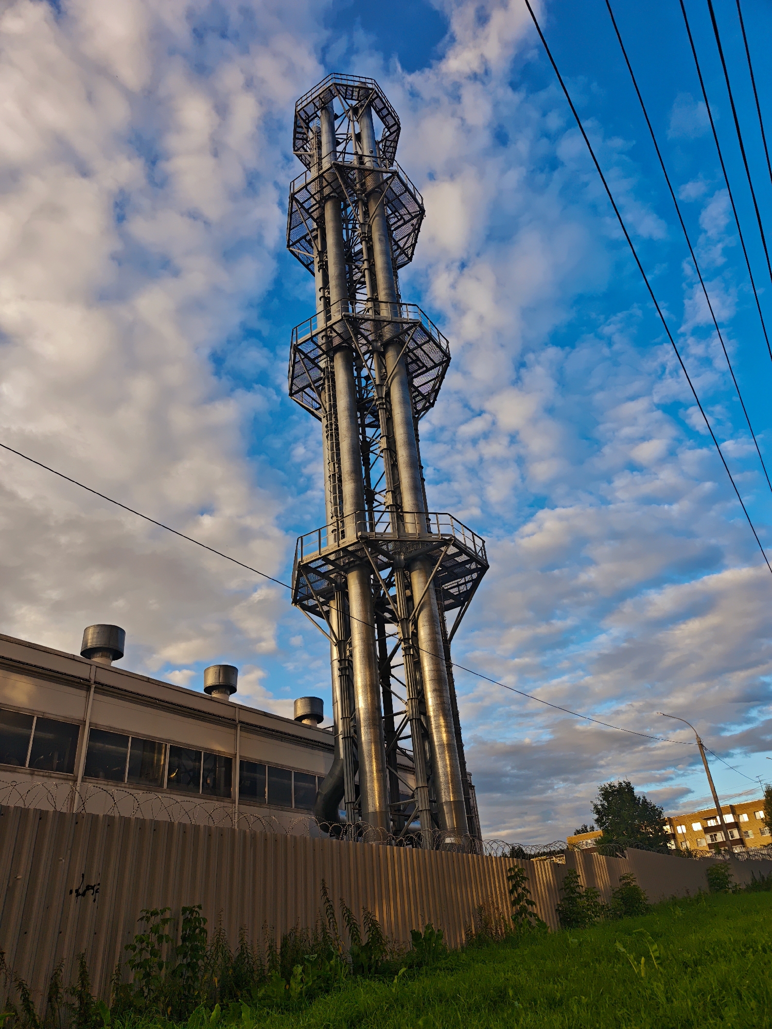 The image depicts an industrial facility in Dmitrov, Russia. In the foreground, a tall, multi-tubed metallic structure, resembling a chimney or exhaust tower, dominates the scene. It is made of interconnected pipes and supported by a metal scaffolding framework with platforms. To the left of this structure is a long, rectangular building with large windows and vents on its roof. A corrugated metal fence, topped with barbed wire, runs across the middle ground, separating the industrial area from a grassy area in the foreground and a street with trees and residential buildings in the background. The sky is a vibrant blue with scattered white and grey clouds, illuminated by what appears to be late afternoon sunlight. Several power lines are visible crossing the upper portion of the image. No people or animals are present in the frame. The overall impression is one of an active industrial zone within a city.